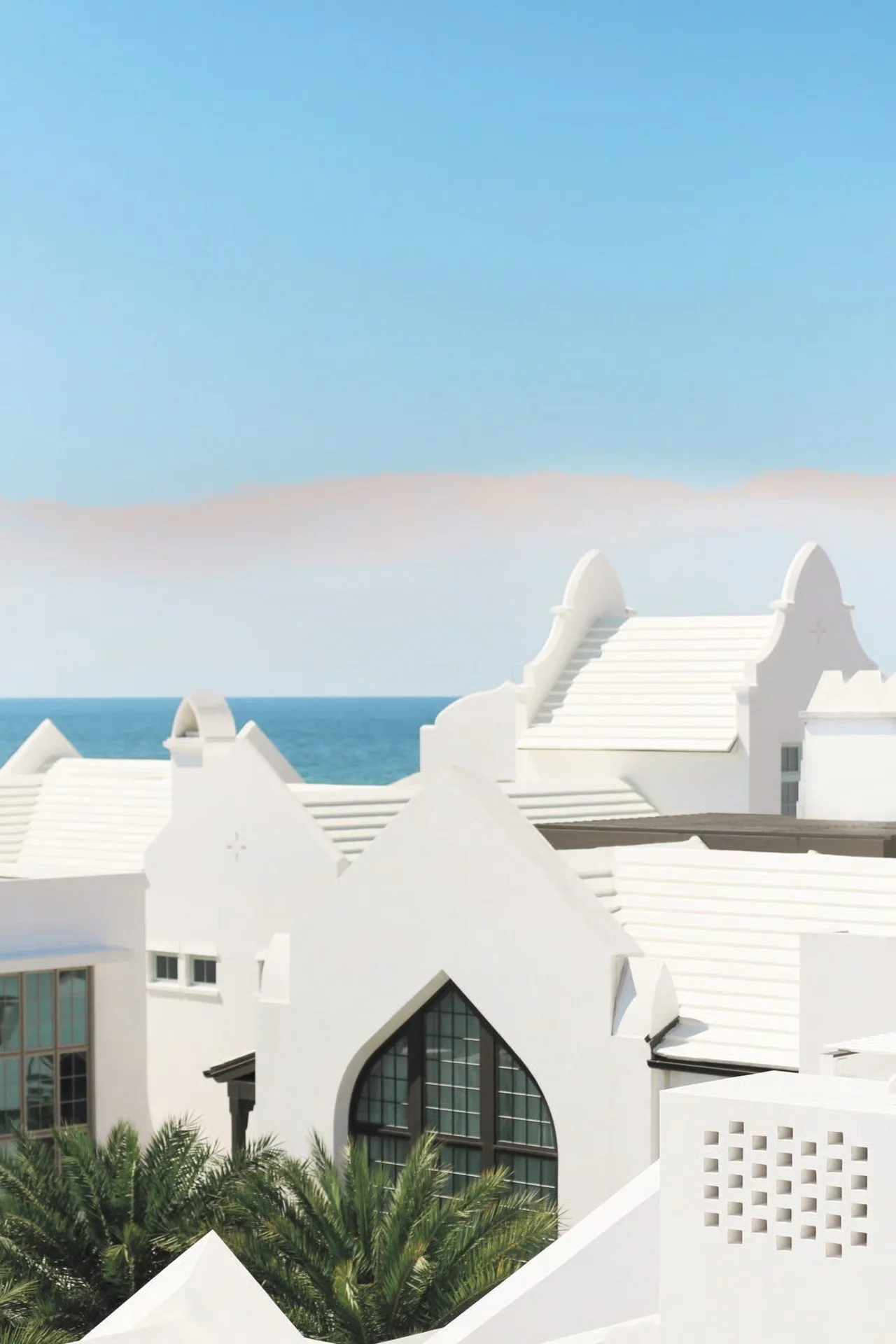 White buildings with modern architectural design by the ocean, featuring curved roofs, large windows, and lush green palm trees in the foreground, under a clear blue sky.