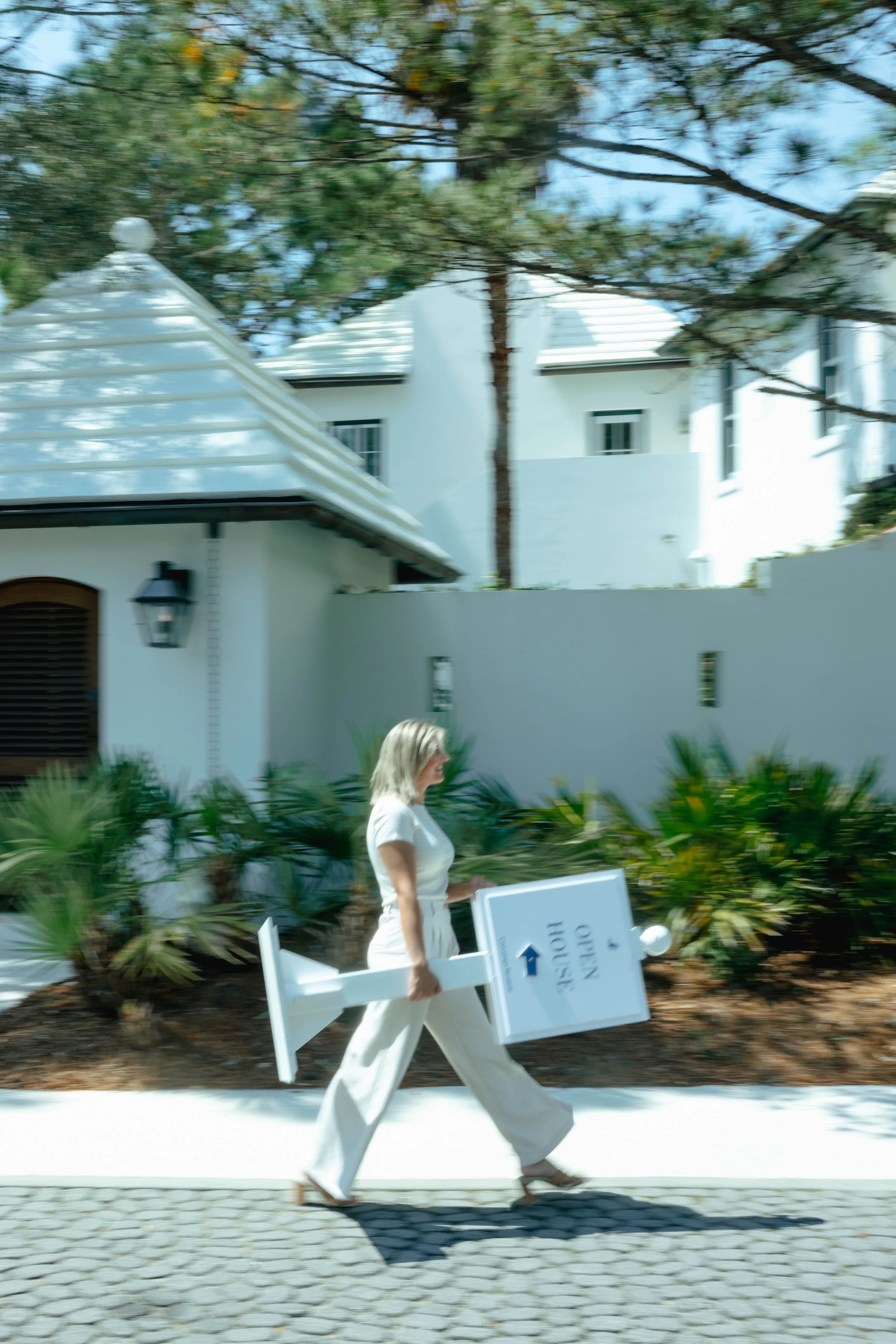 A woman dressed in white walking on a sidewalk while carrying a sign that reads 'VISIT NAPLES.' She is near bushes and appears to be in a residential or hotel area with white buildings and a tree overhead.