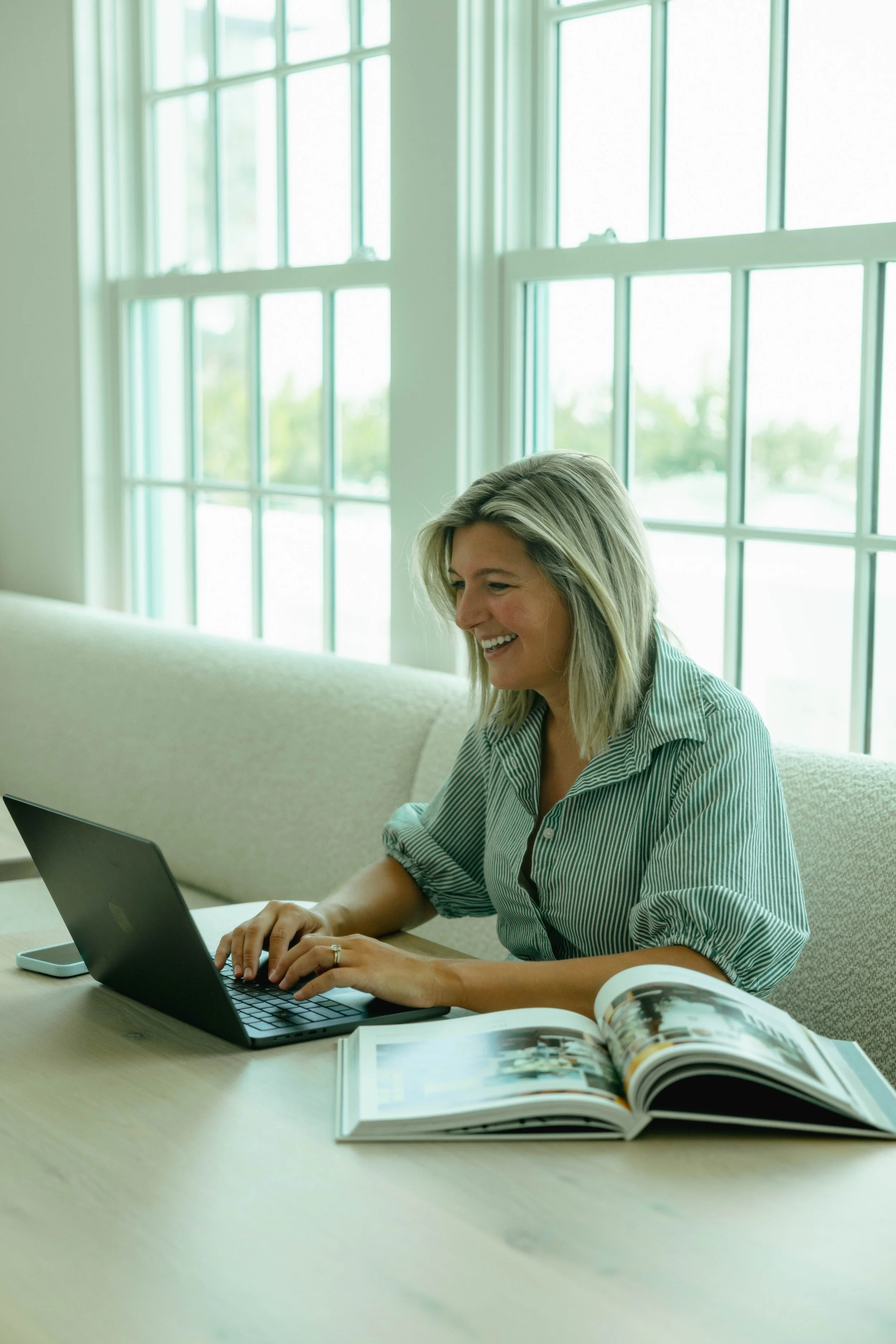 A woman with blonde hair working on a laptop at a light-colored desk in a bright room with large windows. An open magazine is on the desk.