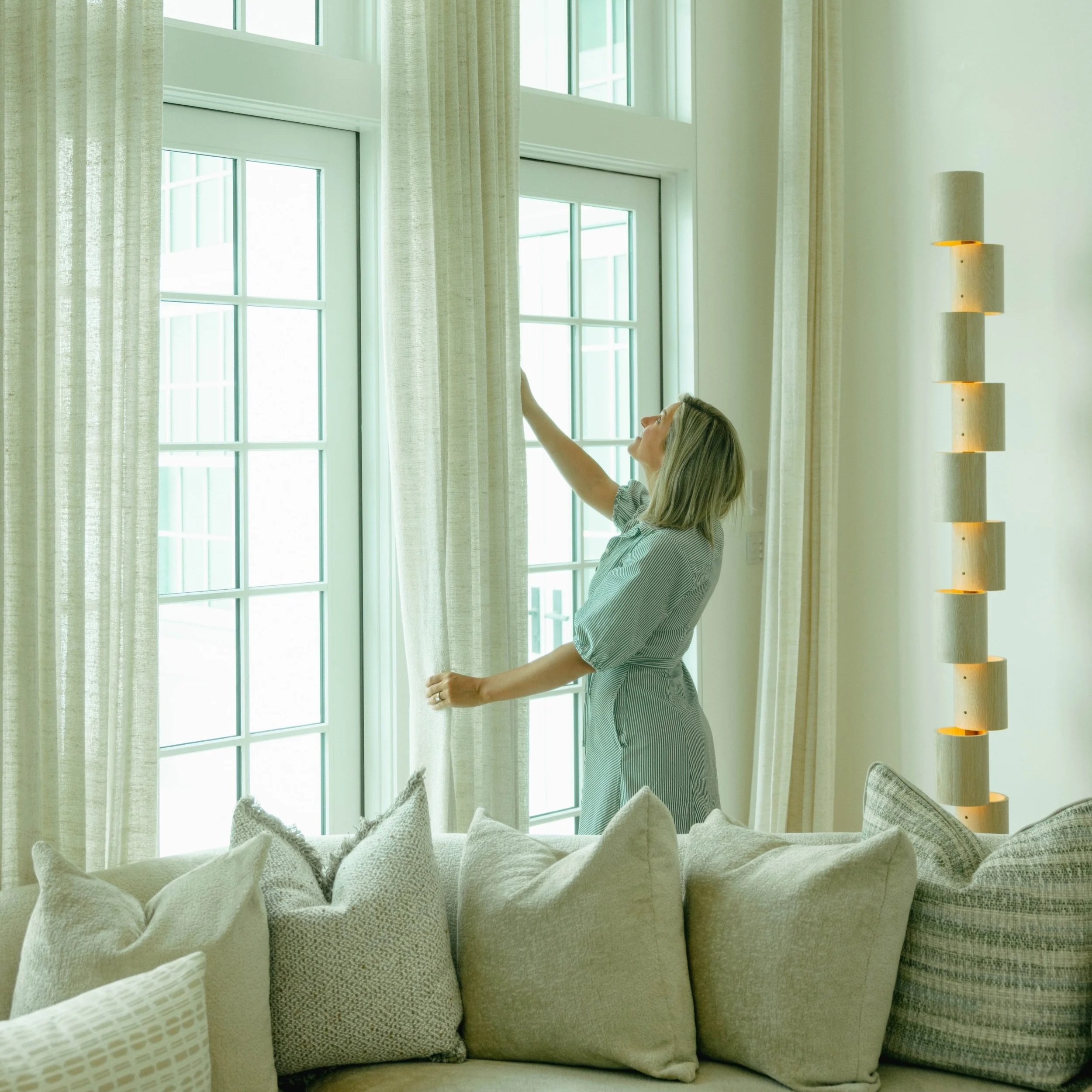 A woman is reaching up to adjust curtains next to a large window, with sheer white curtains hanging beside thicker curtains. In the foreground, there is a light-colored, textured sofa with various cushions.