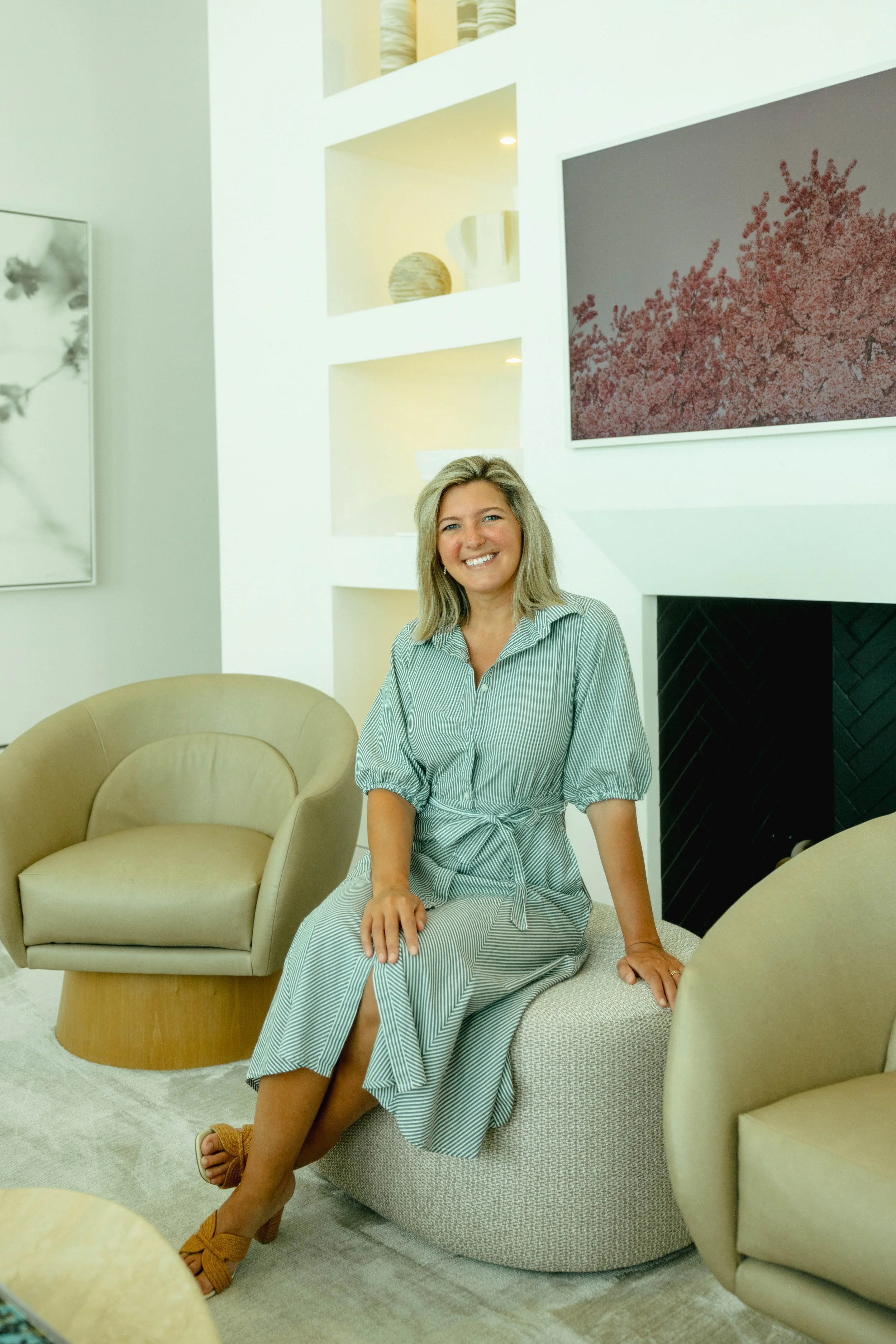 A woman with shoulder-length blonde hair, wearing a light blue and white striped dress, smiling and sitting on a round, textured ottoman in a modern, minimalist living room with cream-colored furniture and abstract artwork on the walls.