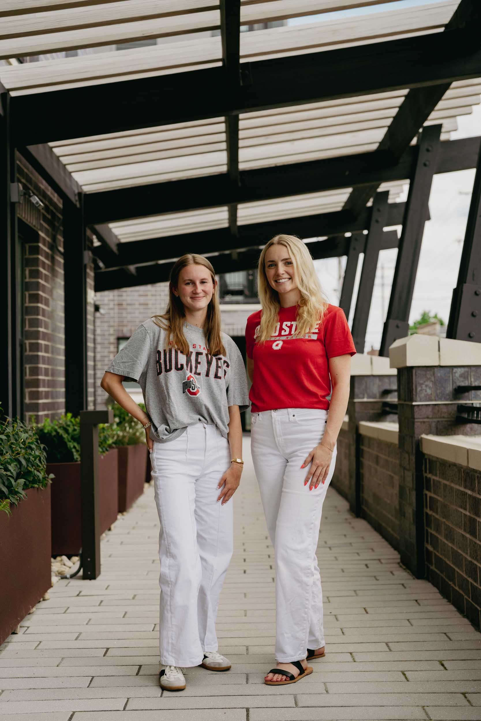 Two women stand on a wooden balcony under a black metal and glass roof, smiling. One wears a gray Ohio State Buckeyes T-shirt and white pants, the other a red Ohio State shirt and white pants. Planters with green plants line the balcony.