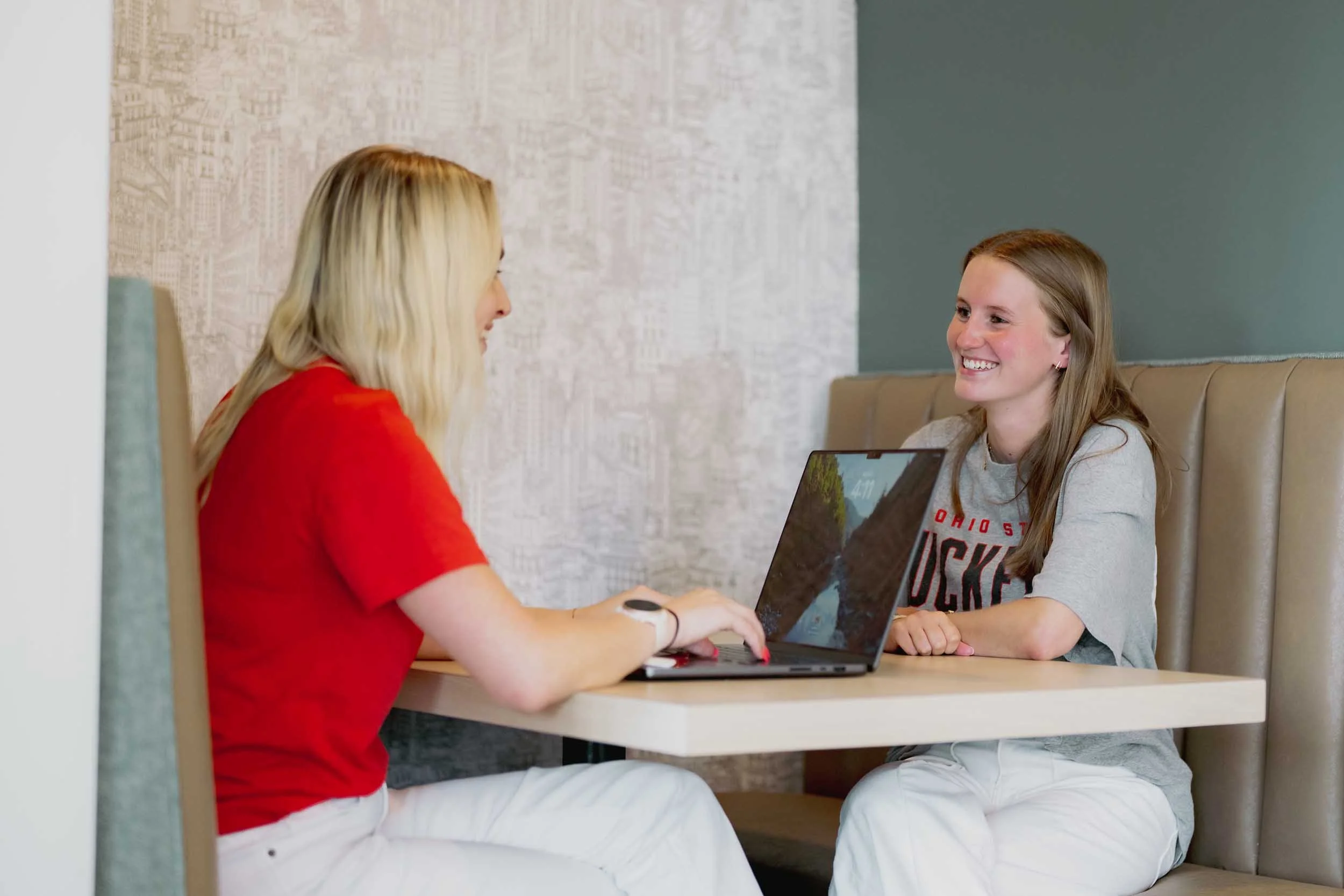 Two women sitting at a table in a cafe, engaging in conversation, one is using a laptop, both smiling. The background features a city map wall art and a dark green wall.