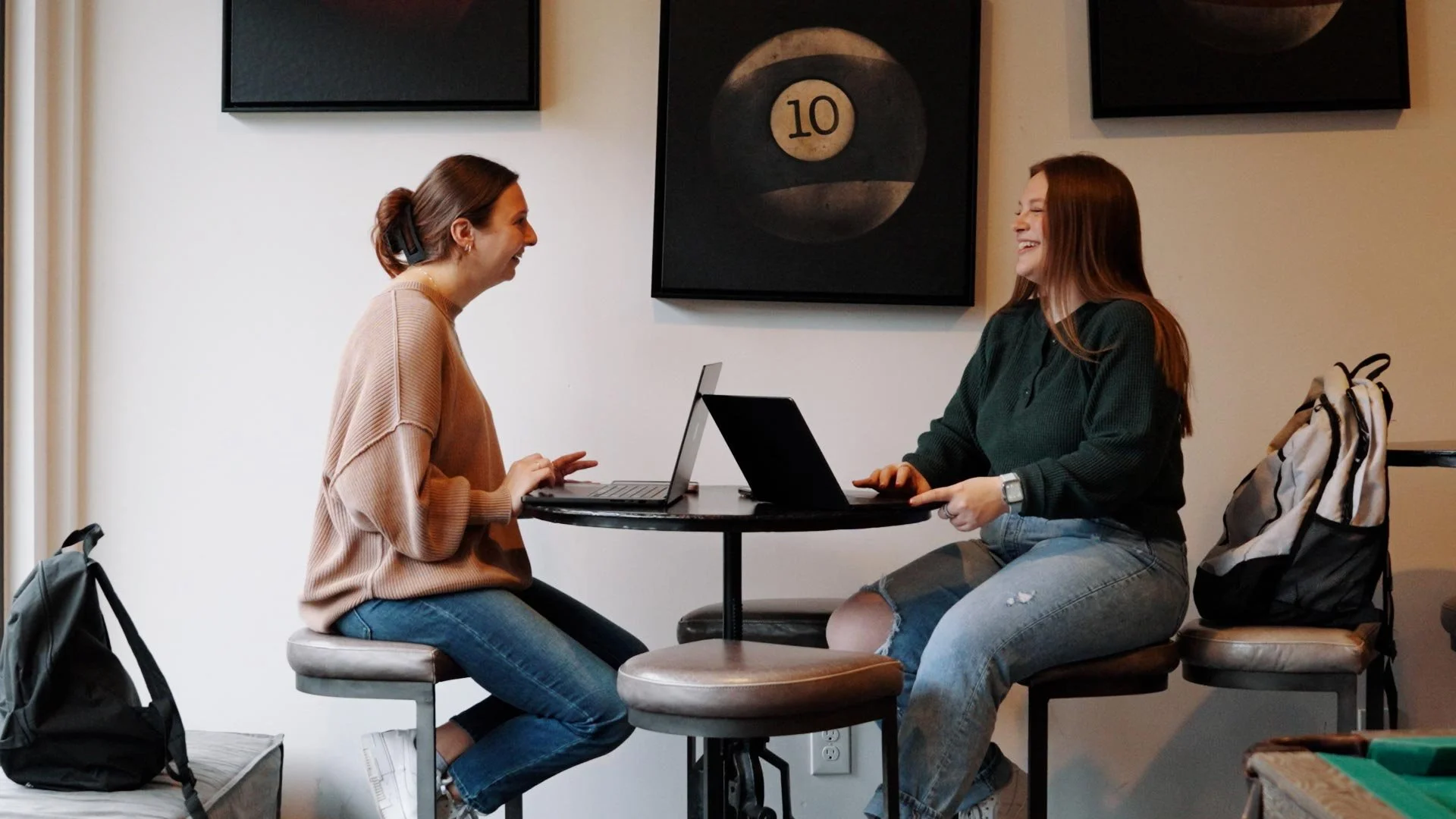 Two women sitting across from each other at a small round table, smiling and talking; each has a laptop open in front of them.