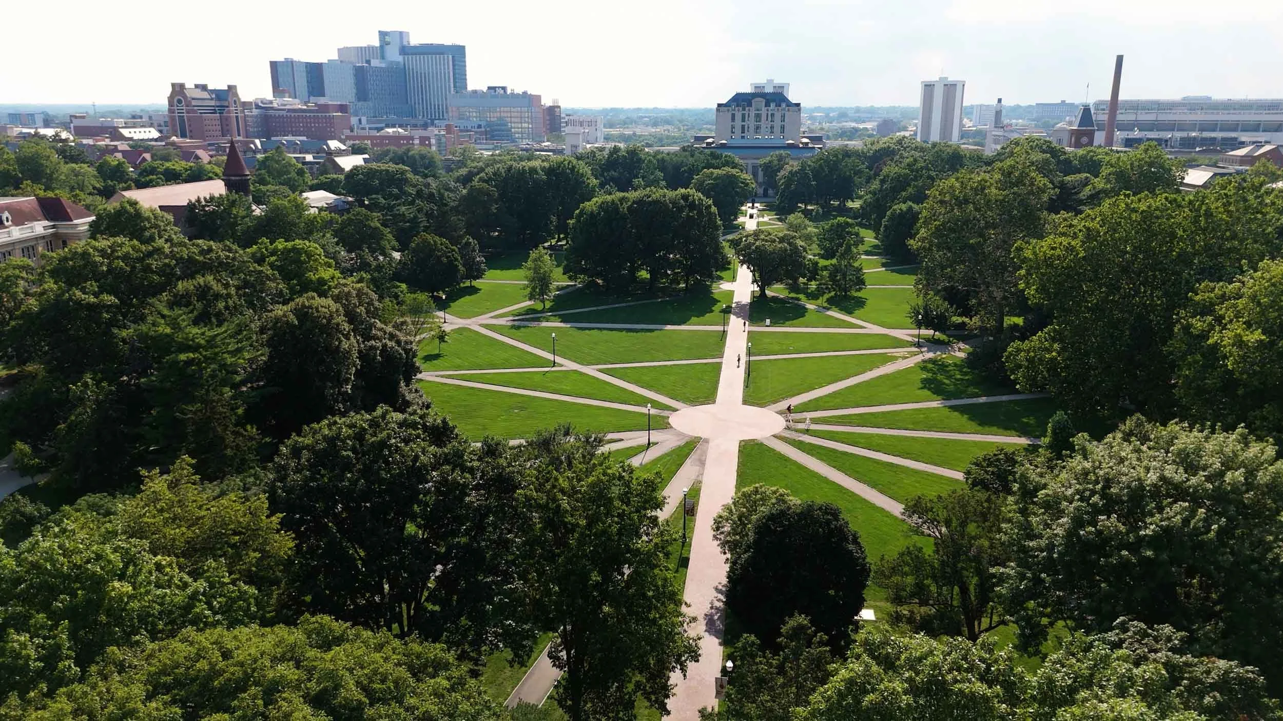 Aerial view of a city park with green trees and grass, intersected by pathways forming a star pattern, with buildings and skyline in the background.