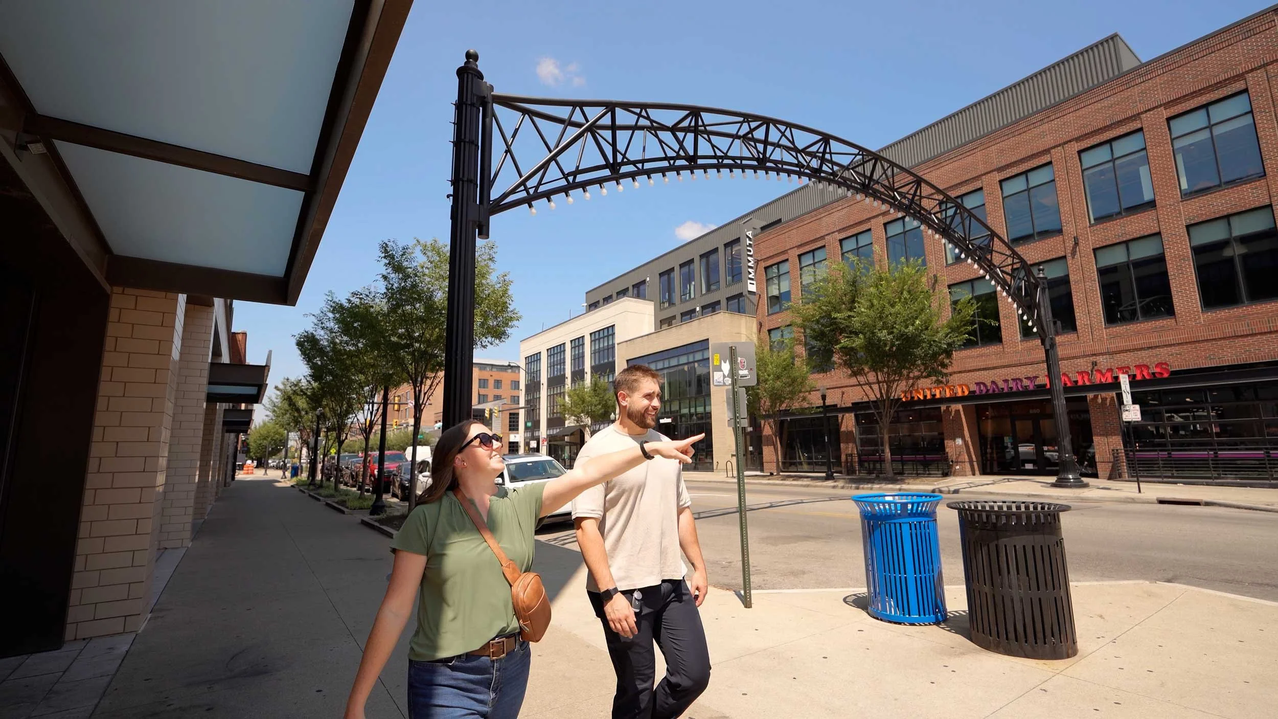 A man and woman walking on a city sidewalk, smiling and pointing, surrounded by buildings, trees, and street furniture on a sunny day.