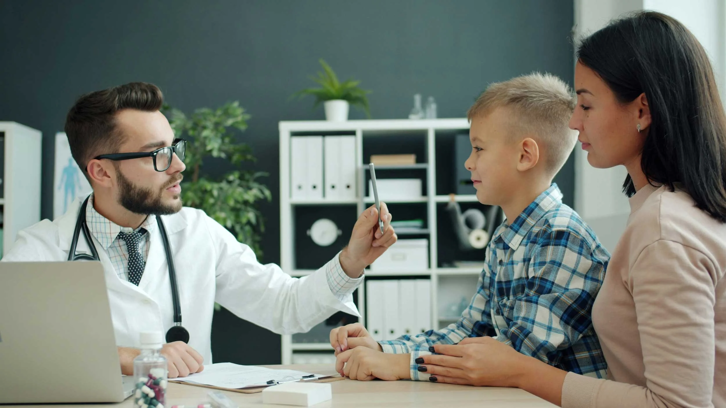A doctor with glasses and a stethoscope around his neck showing a young boy and a woman, likely the child's mother, seated at a table in a medical office.