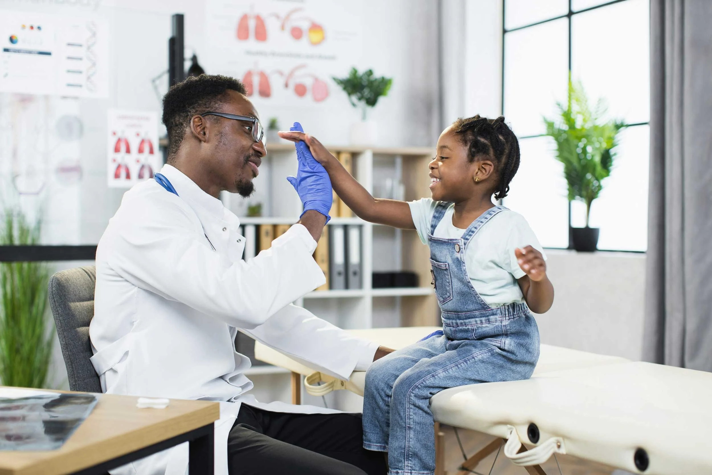 A young girl with braided hair and wearing denim overalls touches foreheads with a male doctor, who is sitting on a chair in a medical office. The doctor is wearing glasses and a white coat. The girl is smiling, and the doctor appears engaged and cheerful. Medical posters and large windows are visible in the background.