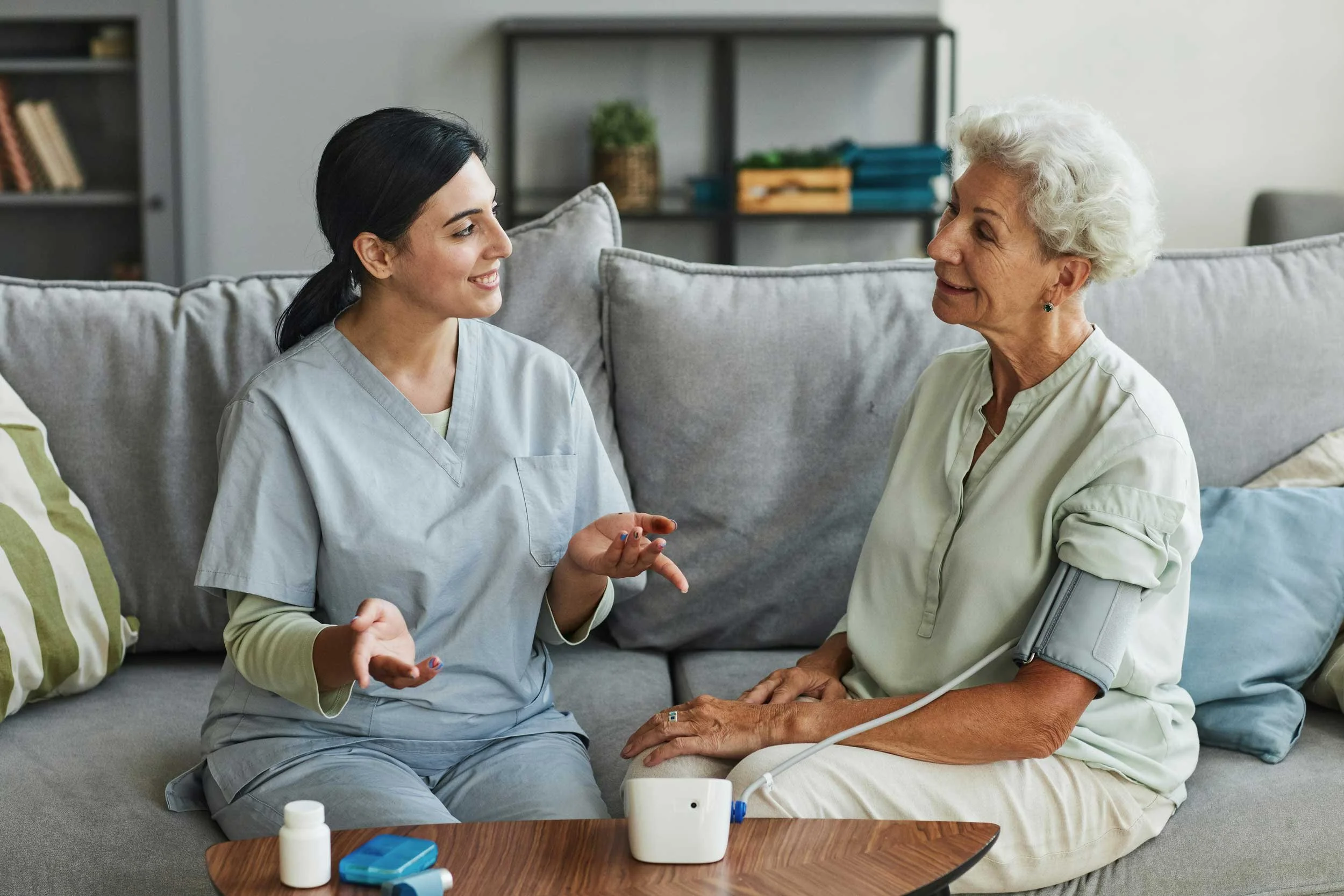A healthcare professional and an elderly woman are sitting on a couch having a conversation, with medical equipment nearby.