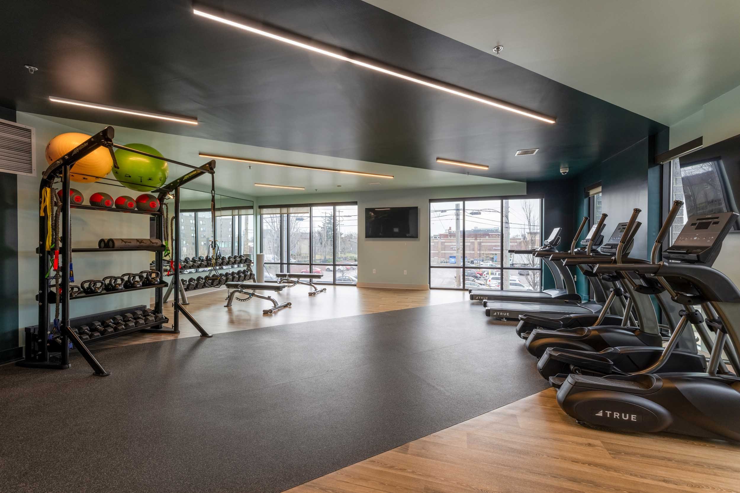 A modern gym with cardio machines, weightlifting equipment, and large windows showing an urban street view.