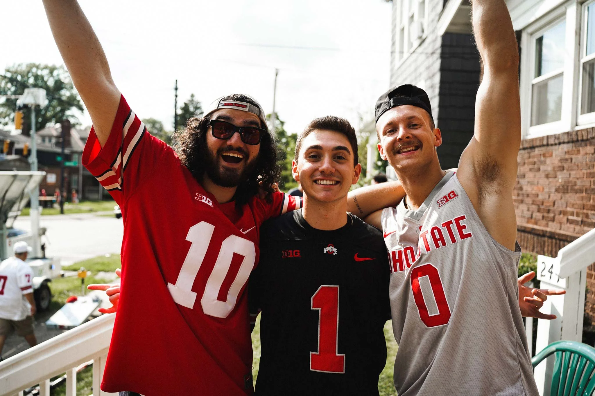 Three young men celebrating together outdoors, smiling with arms around each other. All are wearing Ohio State Buckeyes football jerseys and hats. The background shows a street, some trees, and a building.