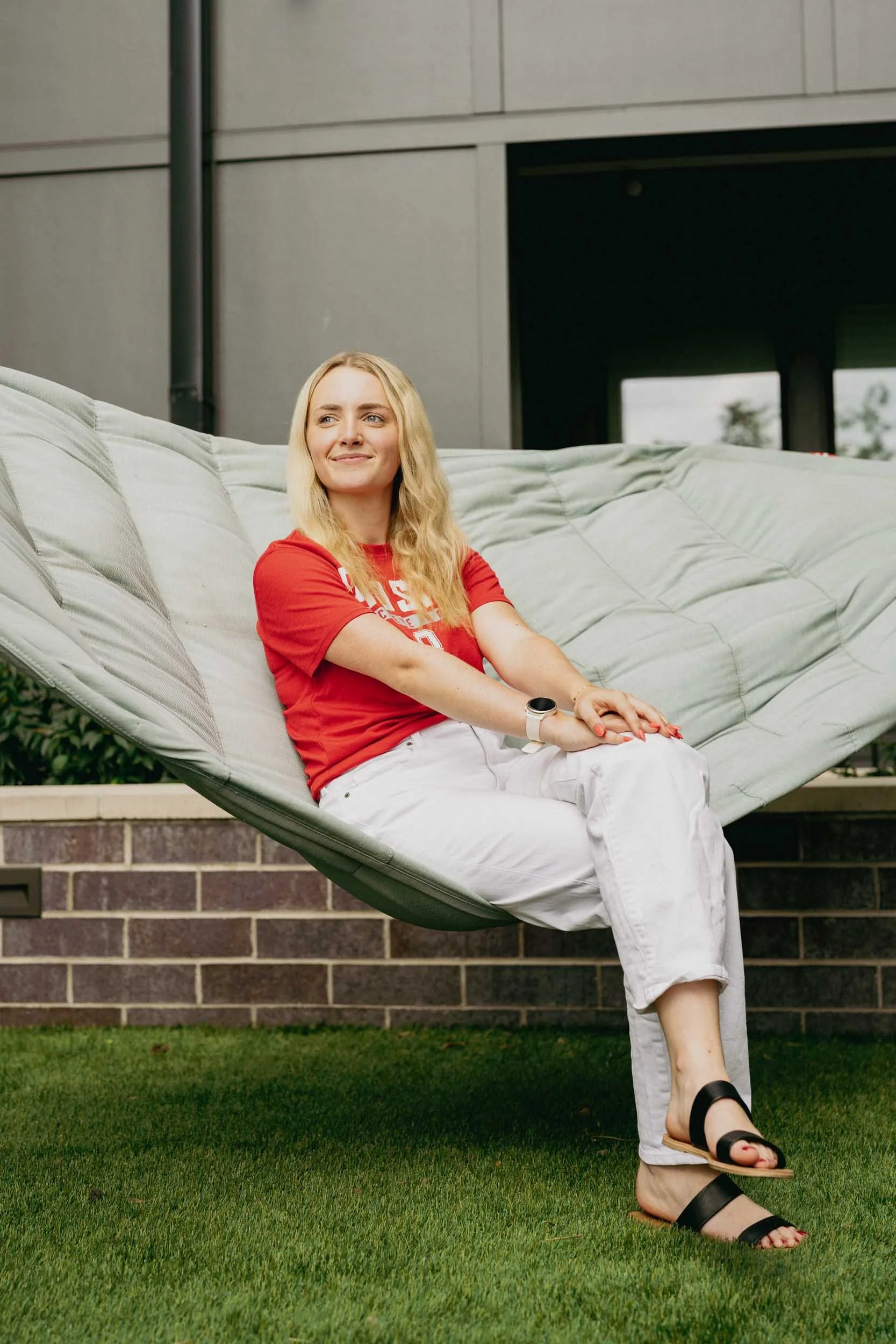 A woman sitting on a large outdoor lounge chair on a grassy area outside a building, smiling and looking to her right, wearing a red T-shirt, white pants, and black sandals.