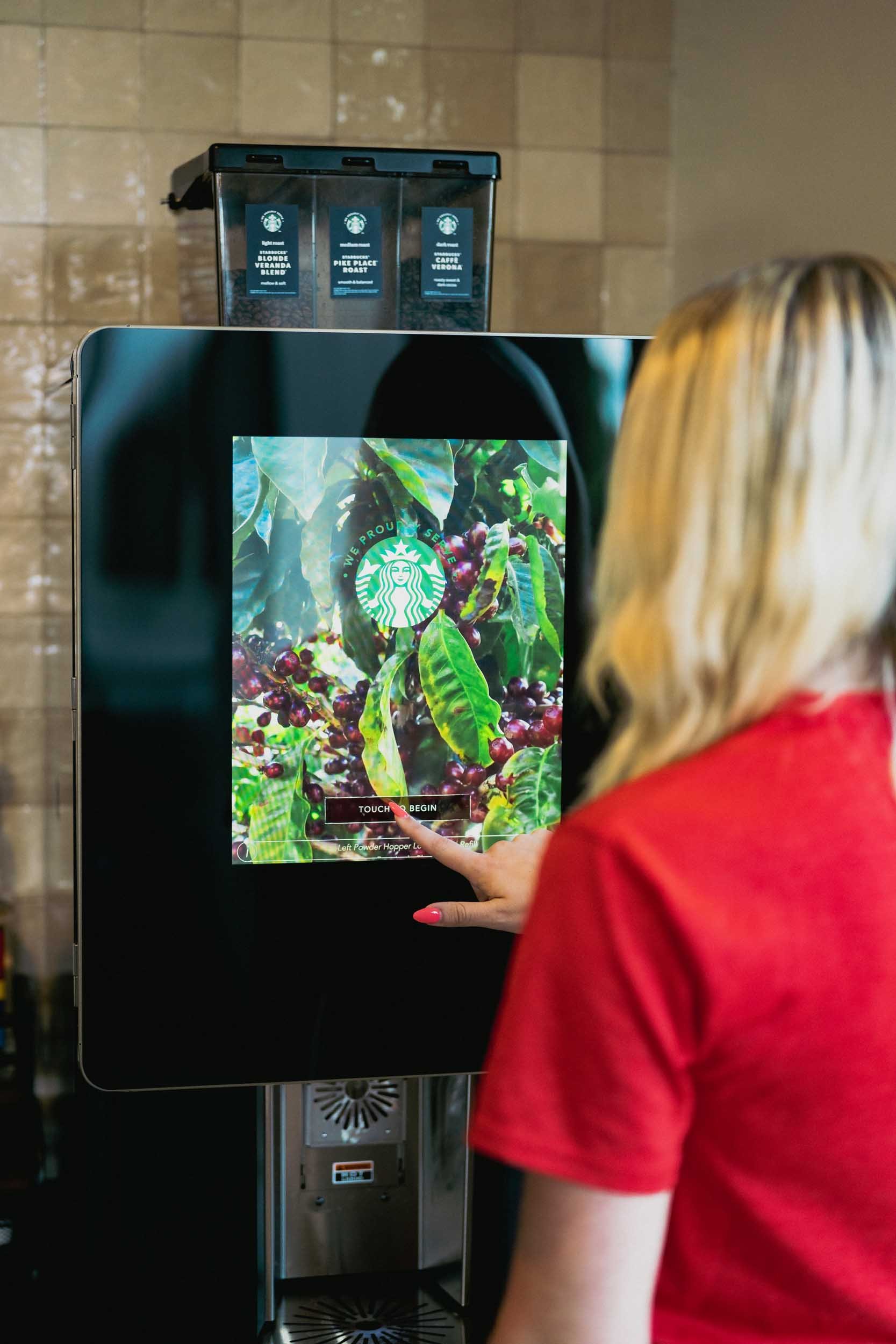 A woman with blonde hair in a red shirt is using a touchscreen coffee ordering kiosk featuring a background of coffee plants and beans, with Starbucks branding and options for coffee selection.