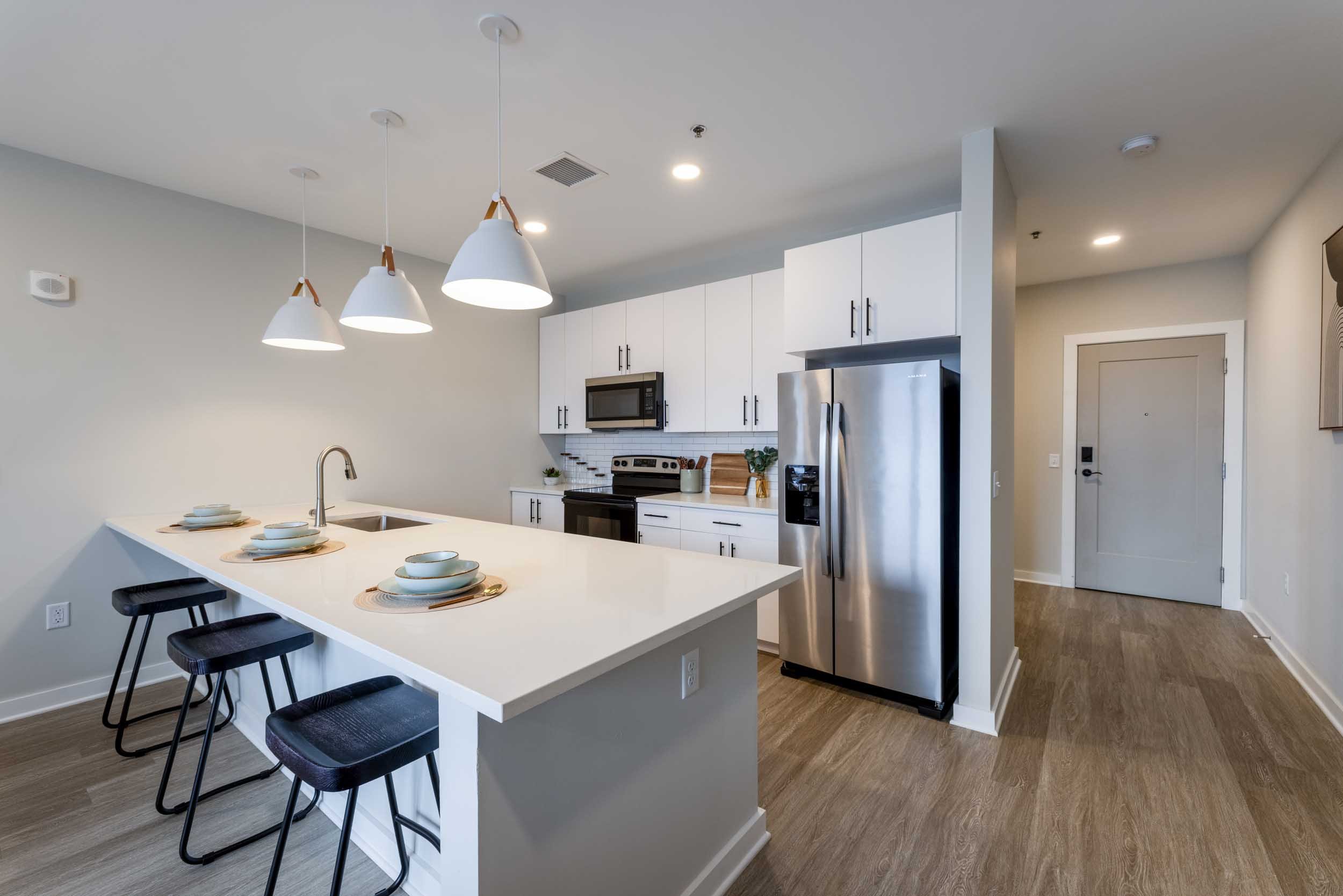Modern kitchen with white island counter, black stools, stainless steel refrigerator, microwave, oven, and white cabinets.