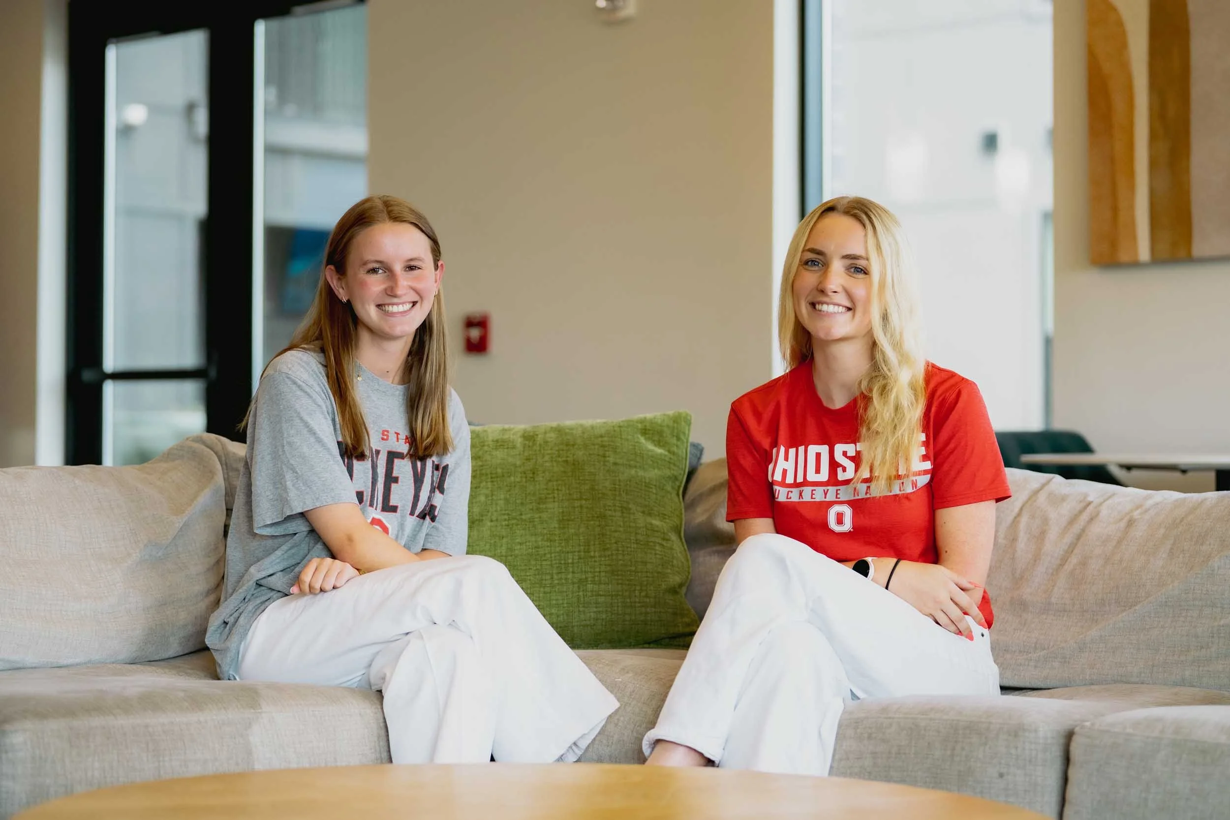 Two young women sitting on a beige couch in a bright room with large windows, smiling at the camera. They are wearing casual t-shirts supporting Ohio State, with one in gray and the other in red, both with white pants.