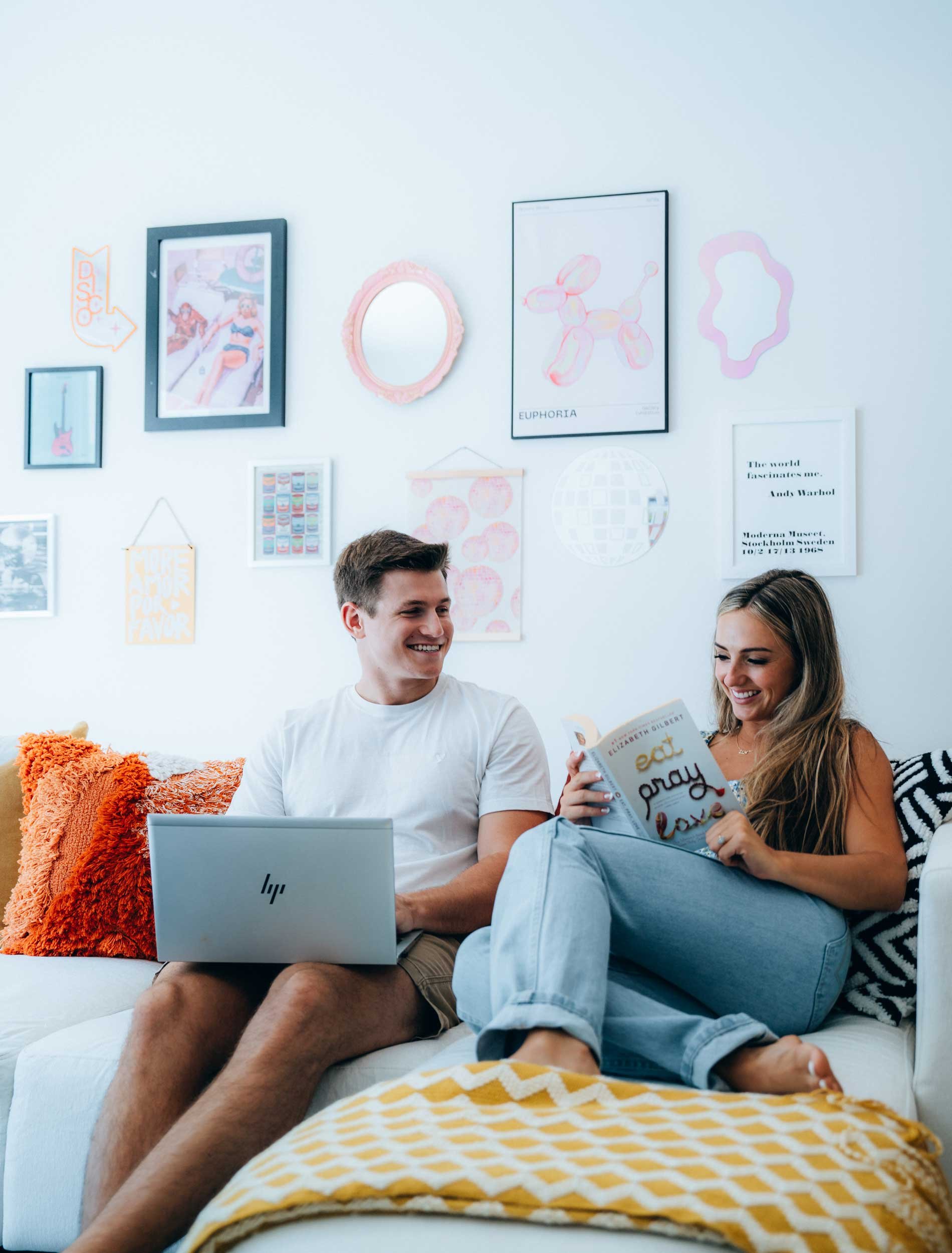 A young man and woman sitting on a sofa, smiling and reading a book together. The room is decorated with framed pictures and wall art, including a balloon dog drawing and a quote. The woman is wearing a striped black and white dress, and they are surrounded by colorful cushions and a yellow and white patterned blanket.