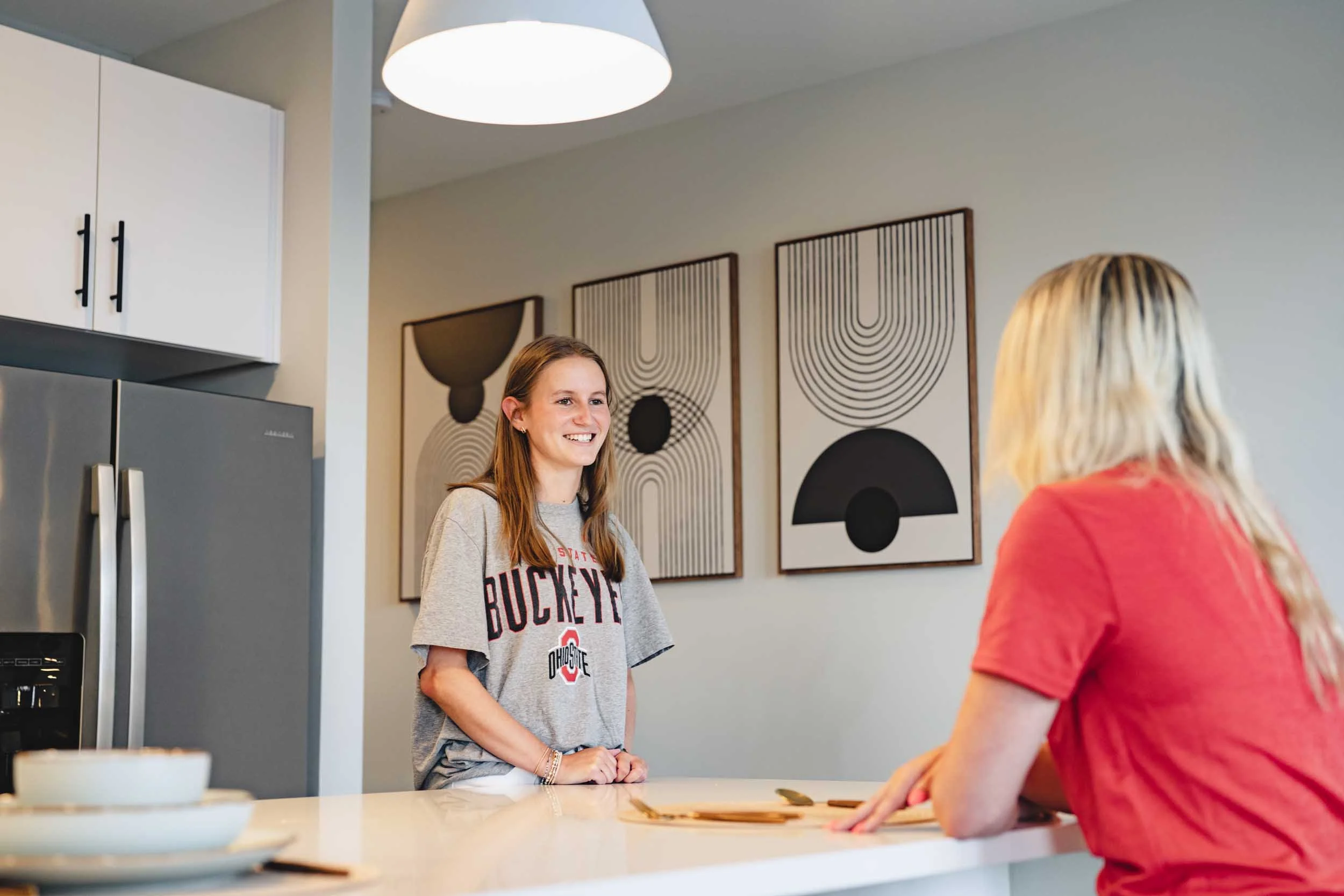 Two women having a conversation in a modern kitchen, with abstract art on the wall, a refrigerator, and a white countertop.