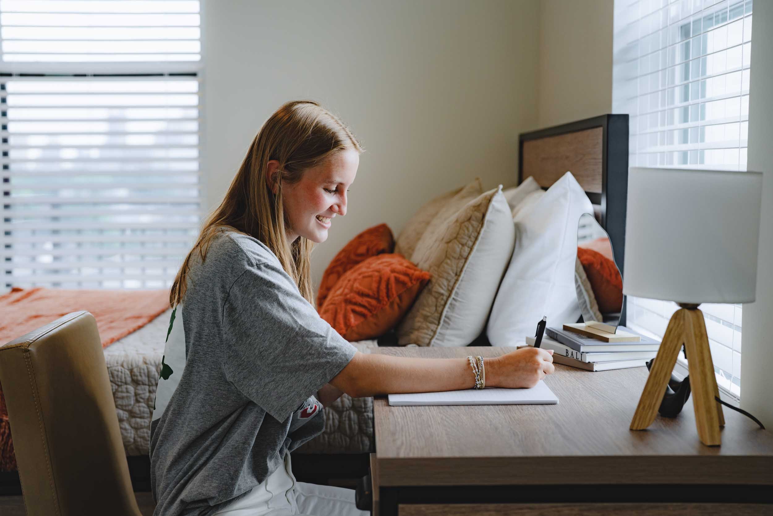 A young girl with long hair sitting at a wooden desk in a well-lit room, smiling and writing in a notebook. There are multiple pillows on the bed behind her and modern window blinds.