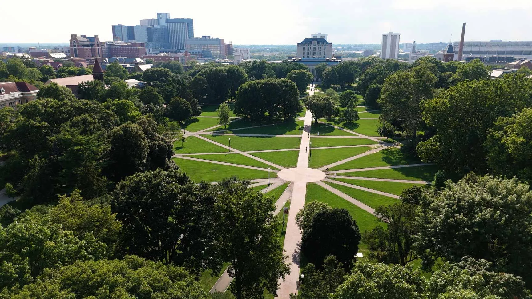 Aerial view of a city park with a circular central area and multiple walking paths radiating outward. Surrounding the park are numerous trees and city buildings in the background.