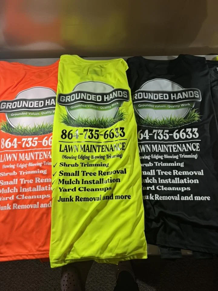 Three yard maintenance service shirts in orange, yellow, and black, with the yellow shirt in the center displaying the company name 'Grounded Hands' and a phone number, along with services such as shrub trimming, small tree removal, mulch installation, yard cleanups, and junk removal.