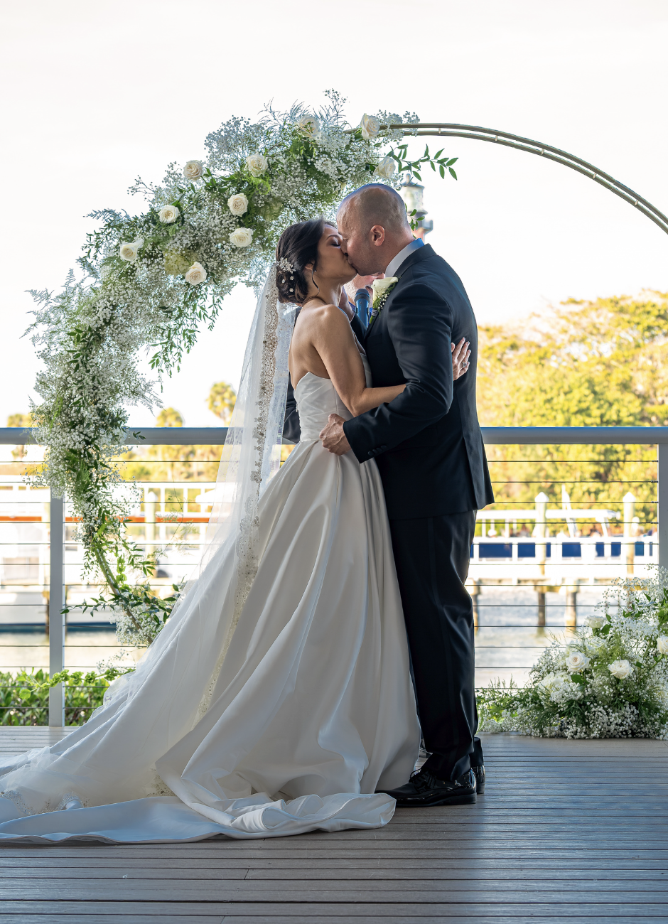 A bride and groom kiss during their wedding ceremony outdoors, standing under a floral arch with white roses and greenery, on a wooden deck with a body of water and trees in the background.