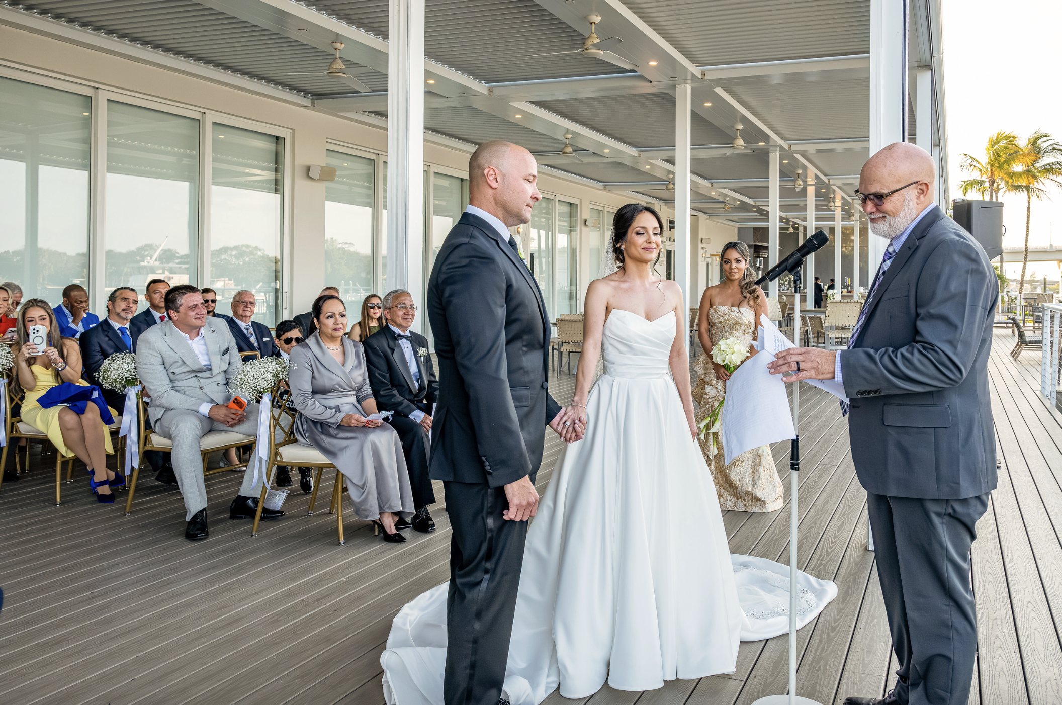 A wedding ceremony taking place outdoors on a deck with a couple standing before an officiant, surrounded by seated guests, some smiling and taking photos, with palm trees and sunlight in the background.