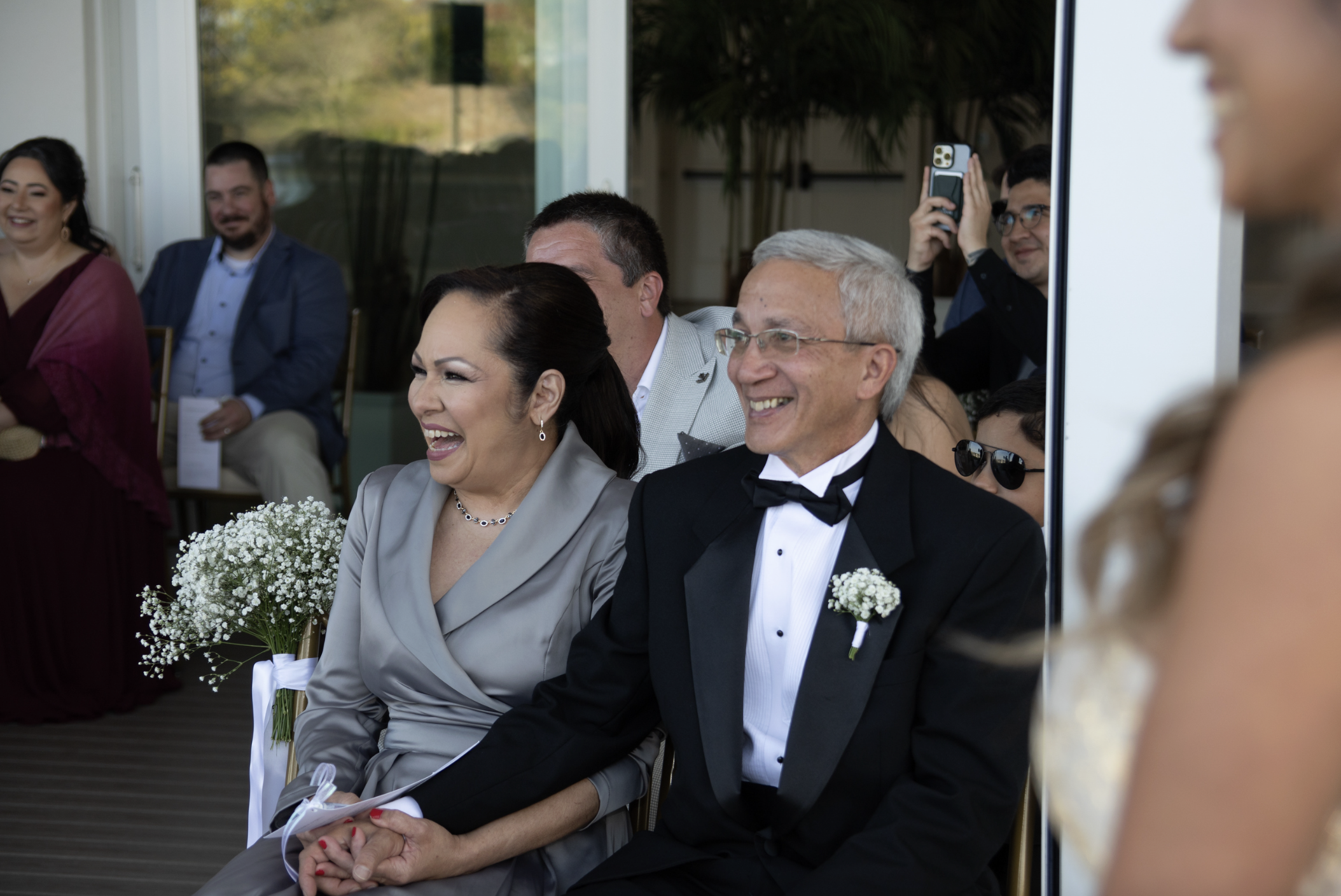 People attending a wedding ceremony, some are smiling and holding bouquets, one person is taking a photo with their phone, everyone dressed in formal attire.