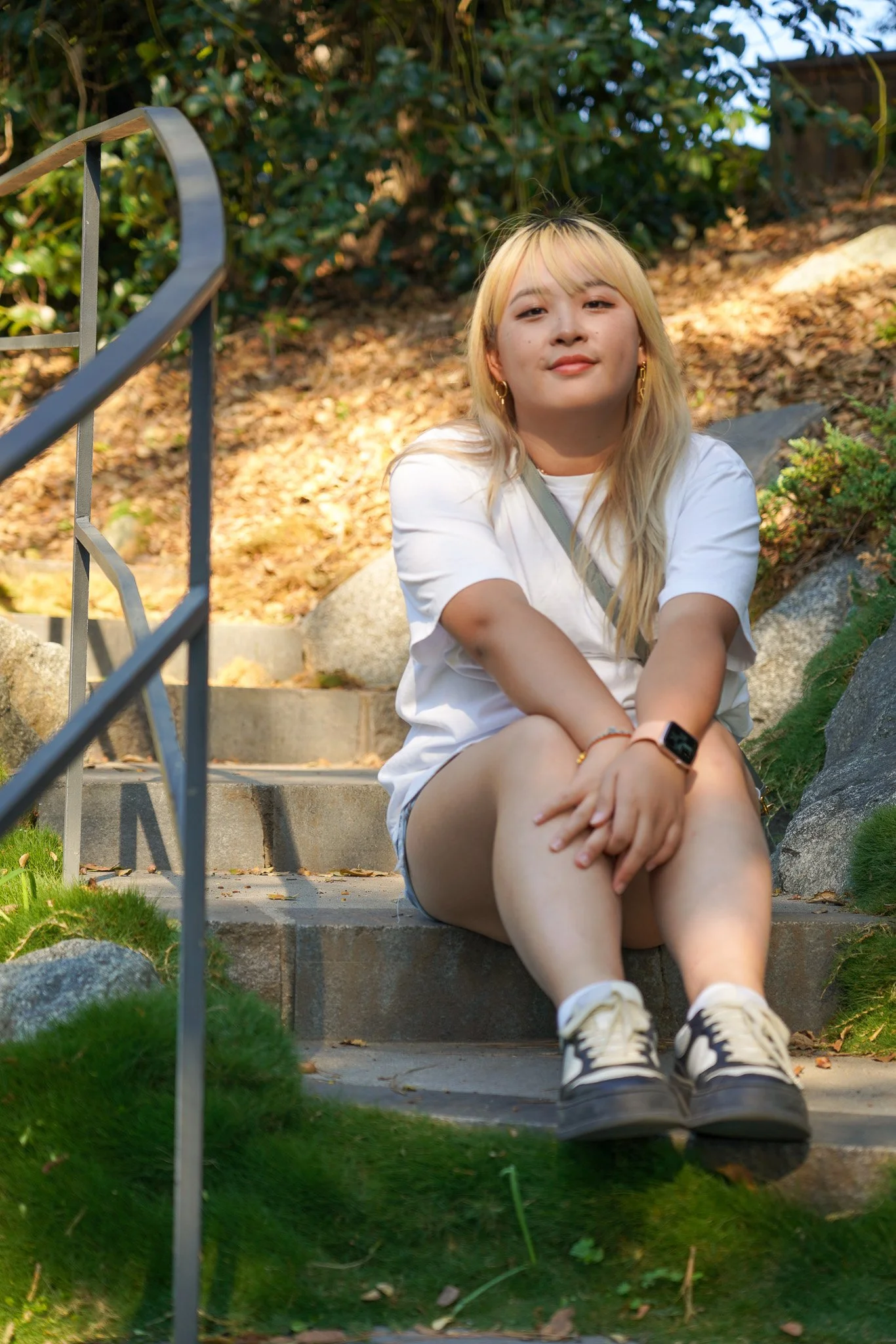 A young woman with blonde hair and gold hoop earrings sitting on outdoor stone stairs, wearing a white t-shirt, shorts, and sneakers, with a relaxed expression, surrounded by greenery and rocks.