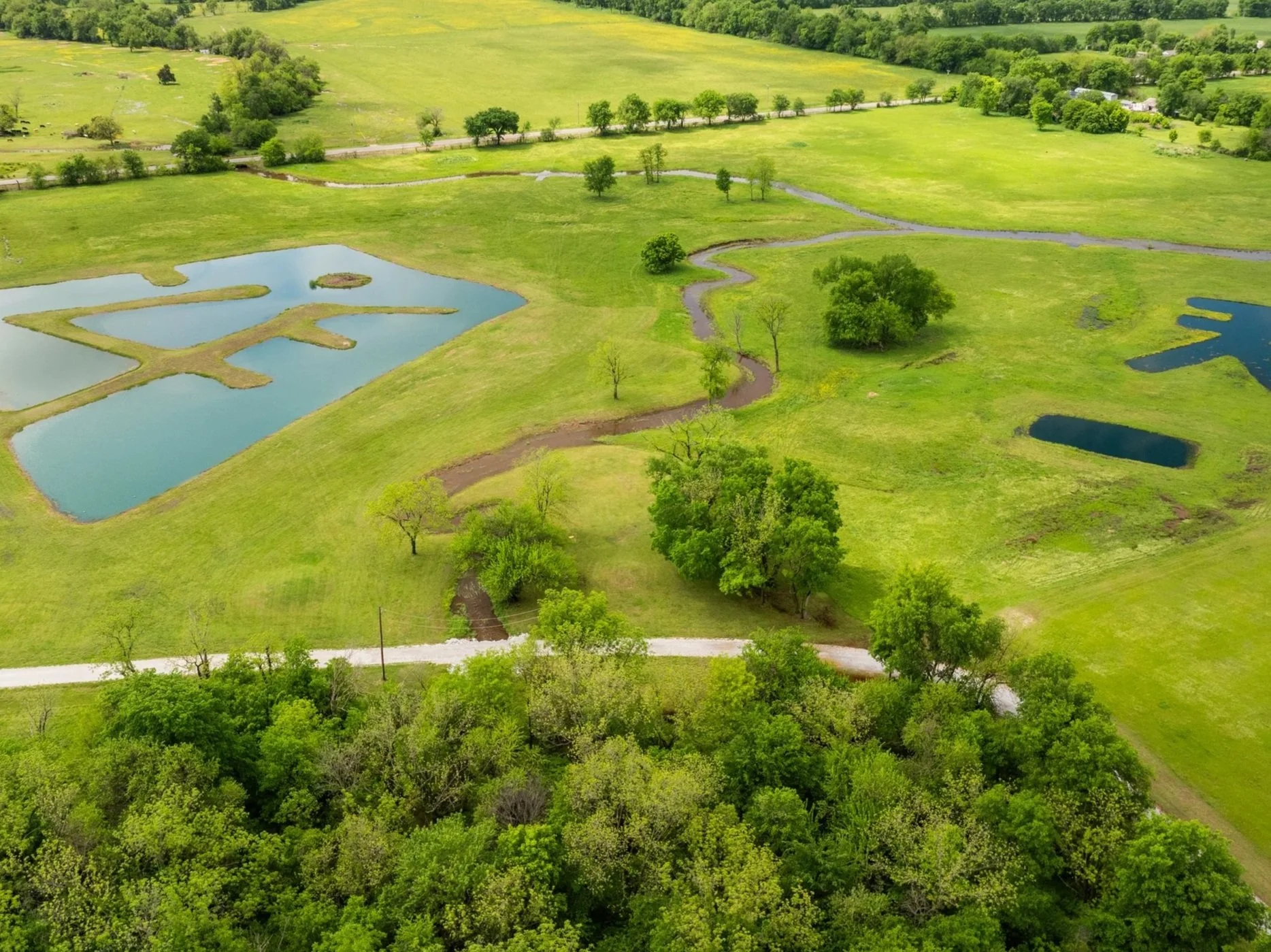 Aerial view of a rural landscape with green fields, small ponds, trees, and a winding dirt road.