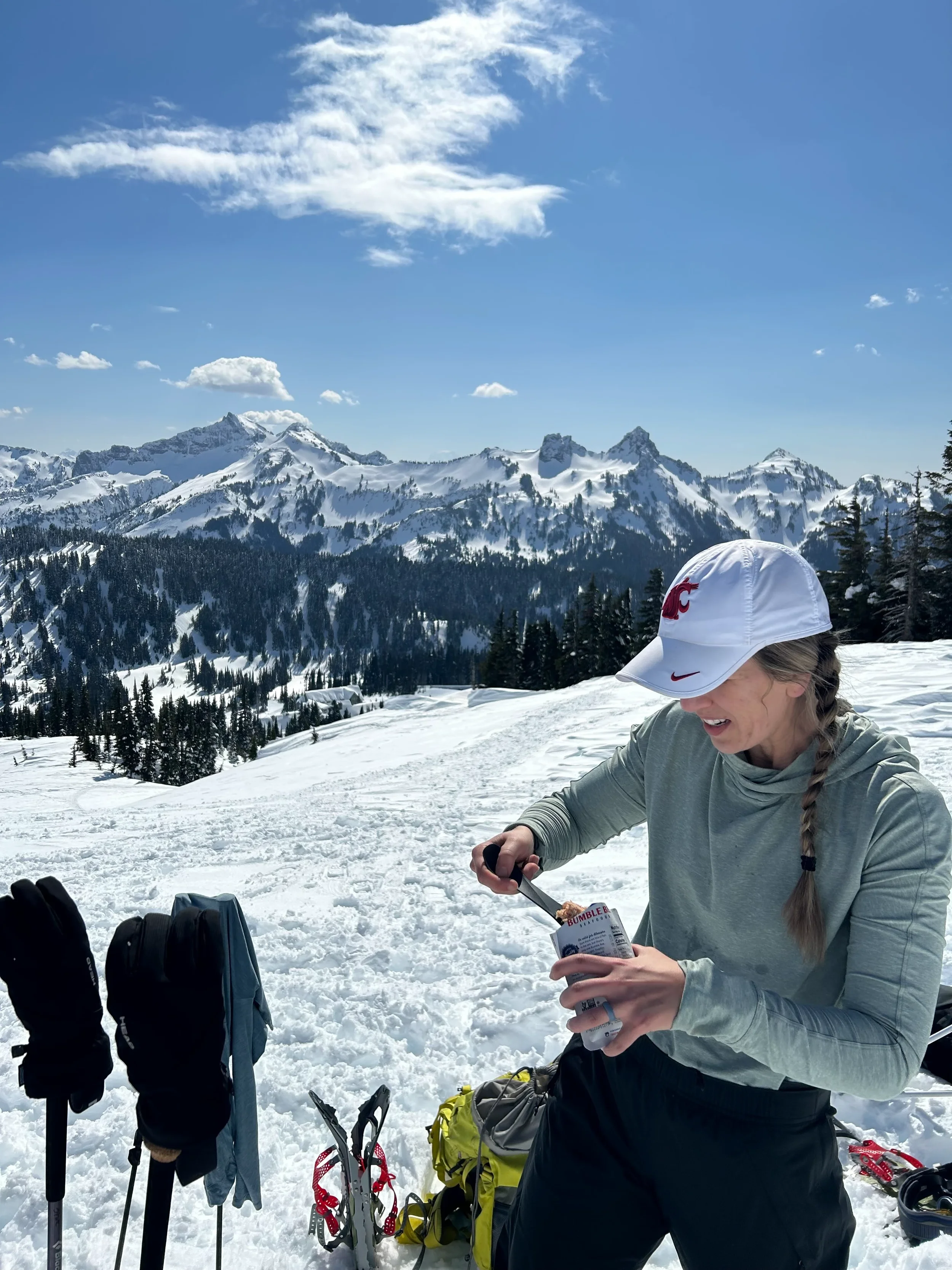 A woman in outdoor winter gear, including a white cap with red logos, is standing on snow with snow-capped mountains and a partly cloudy blue sky in the background. She is opening a snack or food package.