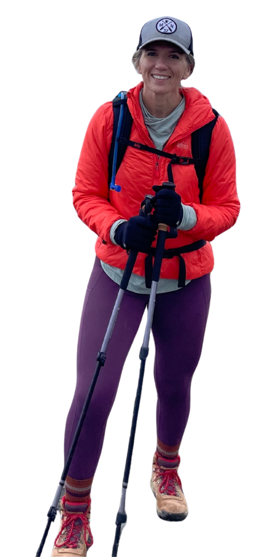 A woman dressed in hiking gear, holding trekking poles, wearing a red jacket, purple pants, hiking boots, and a gray cap with a logo, smiling on a hiking trail.