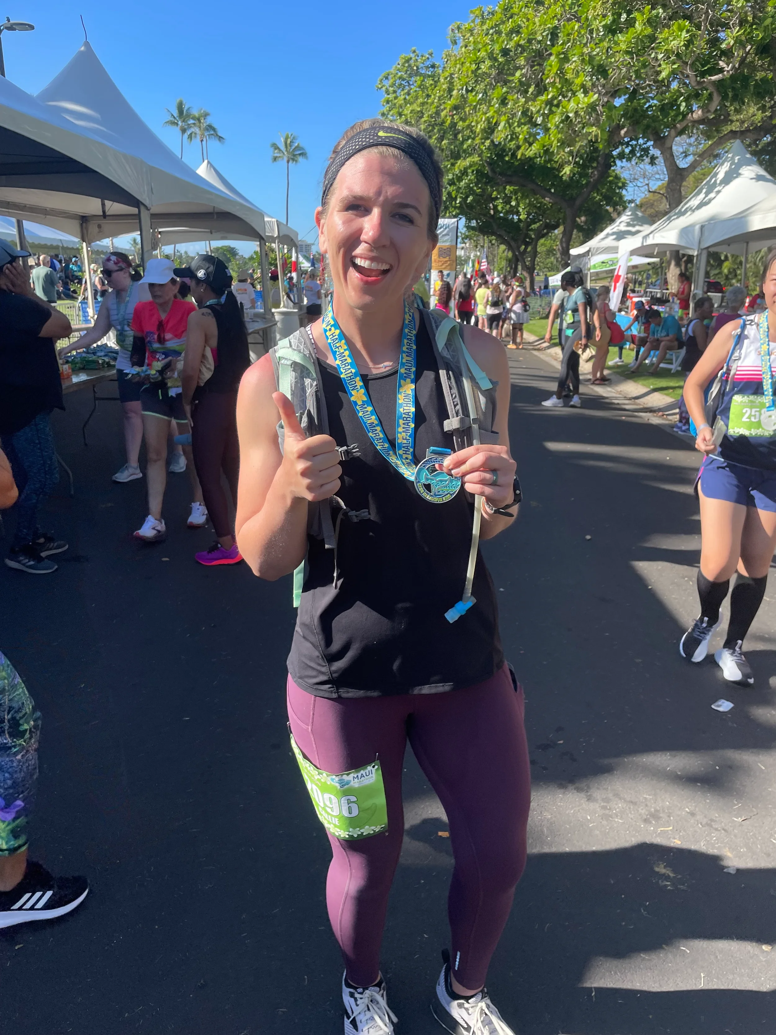 A smiling woman wearing a black sleeveless shirt and purple leggings, giving a thumbs-up at a marathon event with other participants and volunteers under white tents on a sunny day with palm trees and green trees in the background.