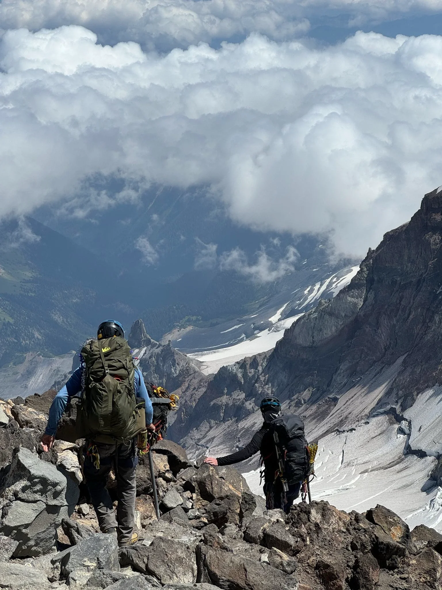 A group of climbers ascending a rocky mountain trail, with snow-covered peaks and clouds in the background.