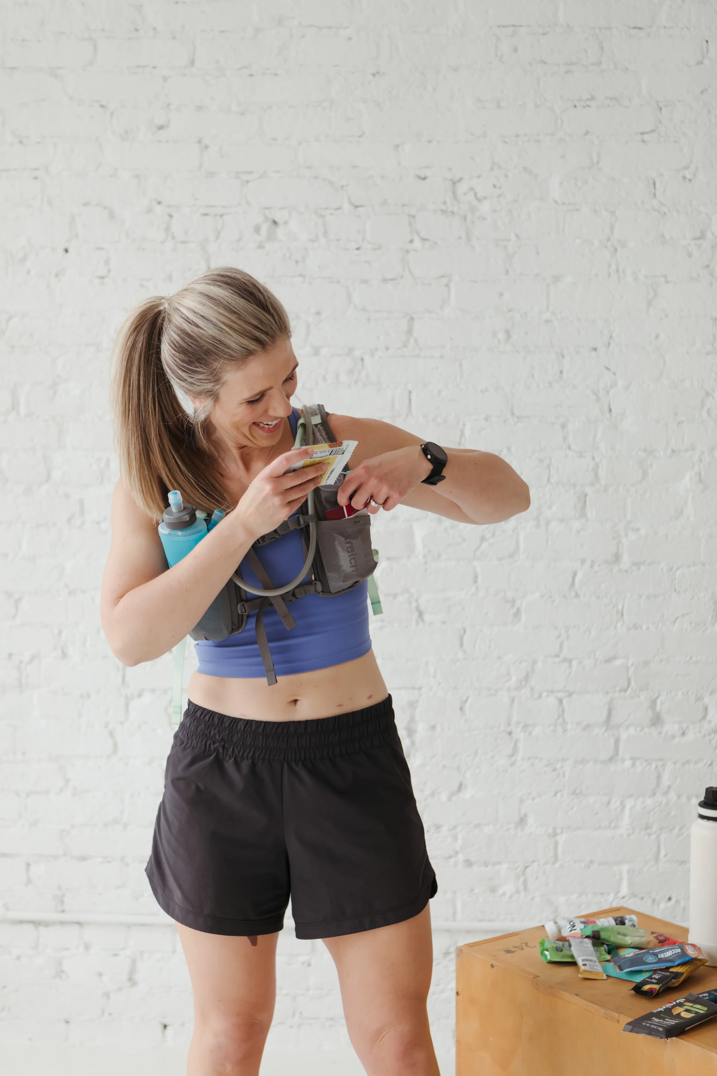 A woman with blonde hair tied back, wearing a blue sports bra and black shorts, examining a snack or food wrapper, with a water bottle attached to her sports gear, standing in a room with a white brick wall. There are various snack packages on a wooden table beside her.