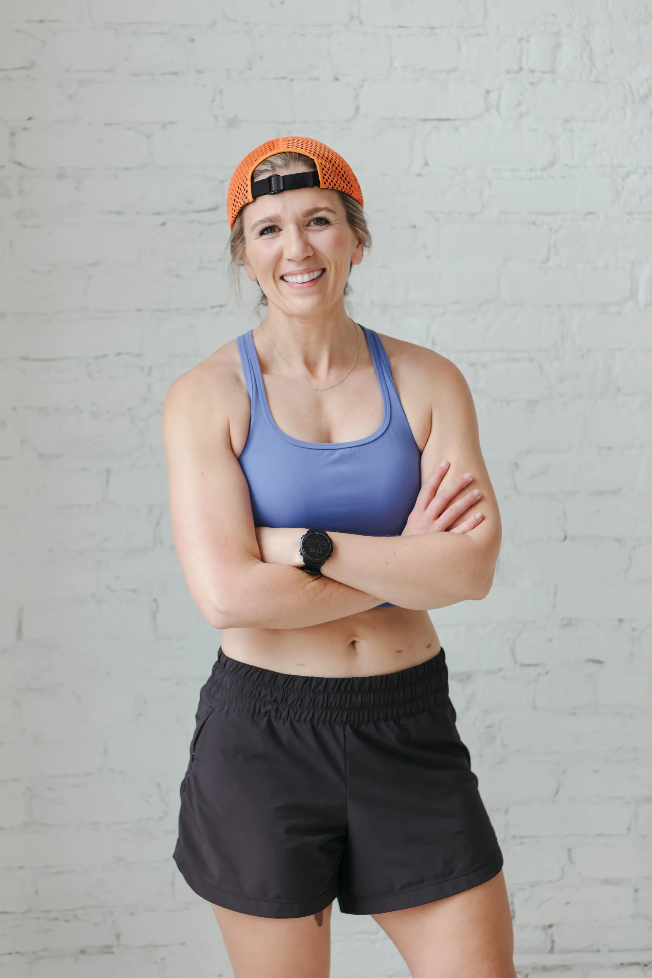 A woman with a smiling expression, wearing an orange mesh baseball cap backwards, a blue sports bra, black athletic shorts, a black wristwatch, standing against a white brick wall with crossed arms.