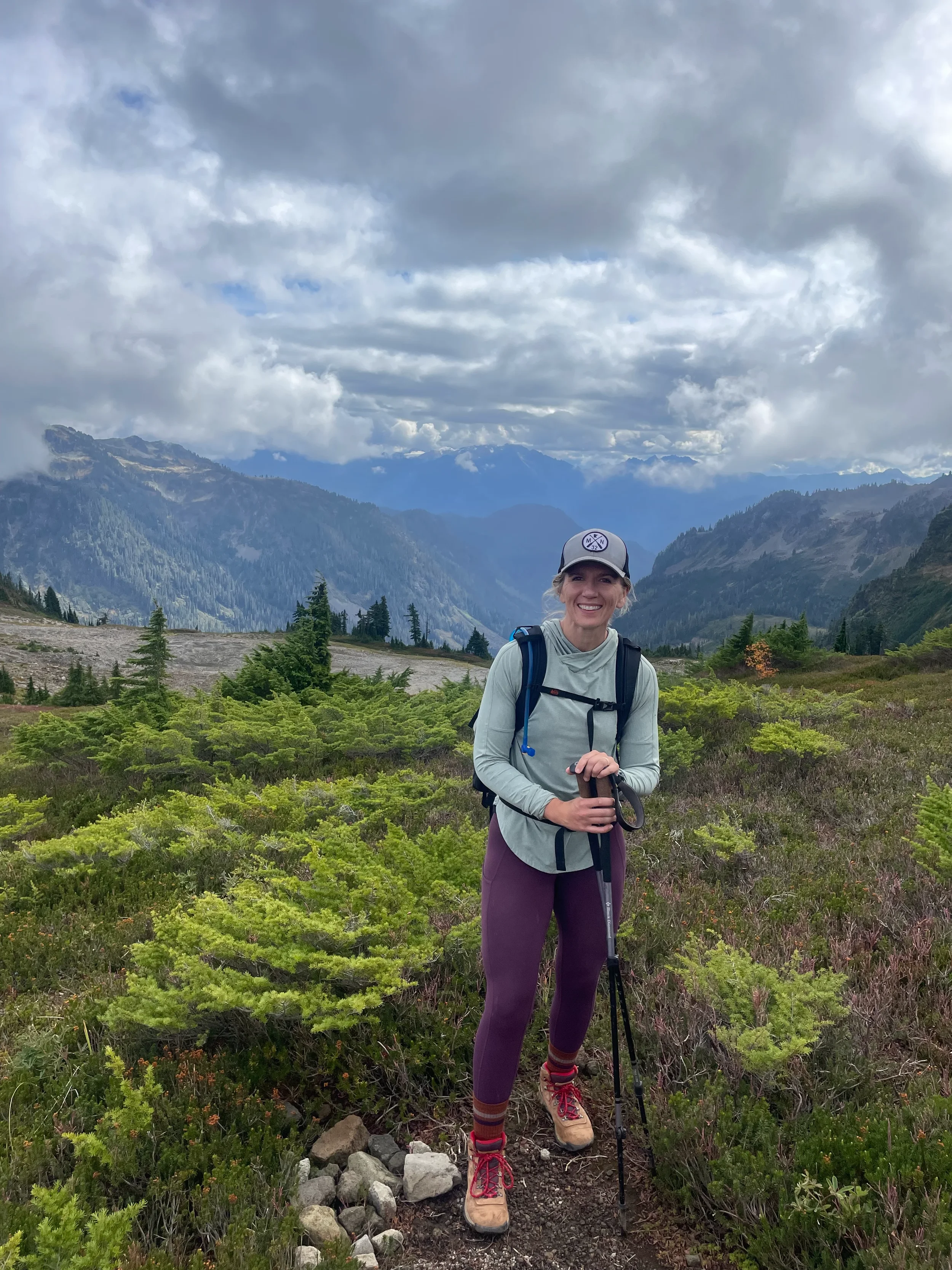 A woman smiling while hiking in a mountainous landscape with lush green vegetation, trees, and a cloudy sky.