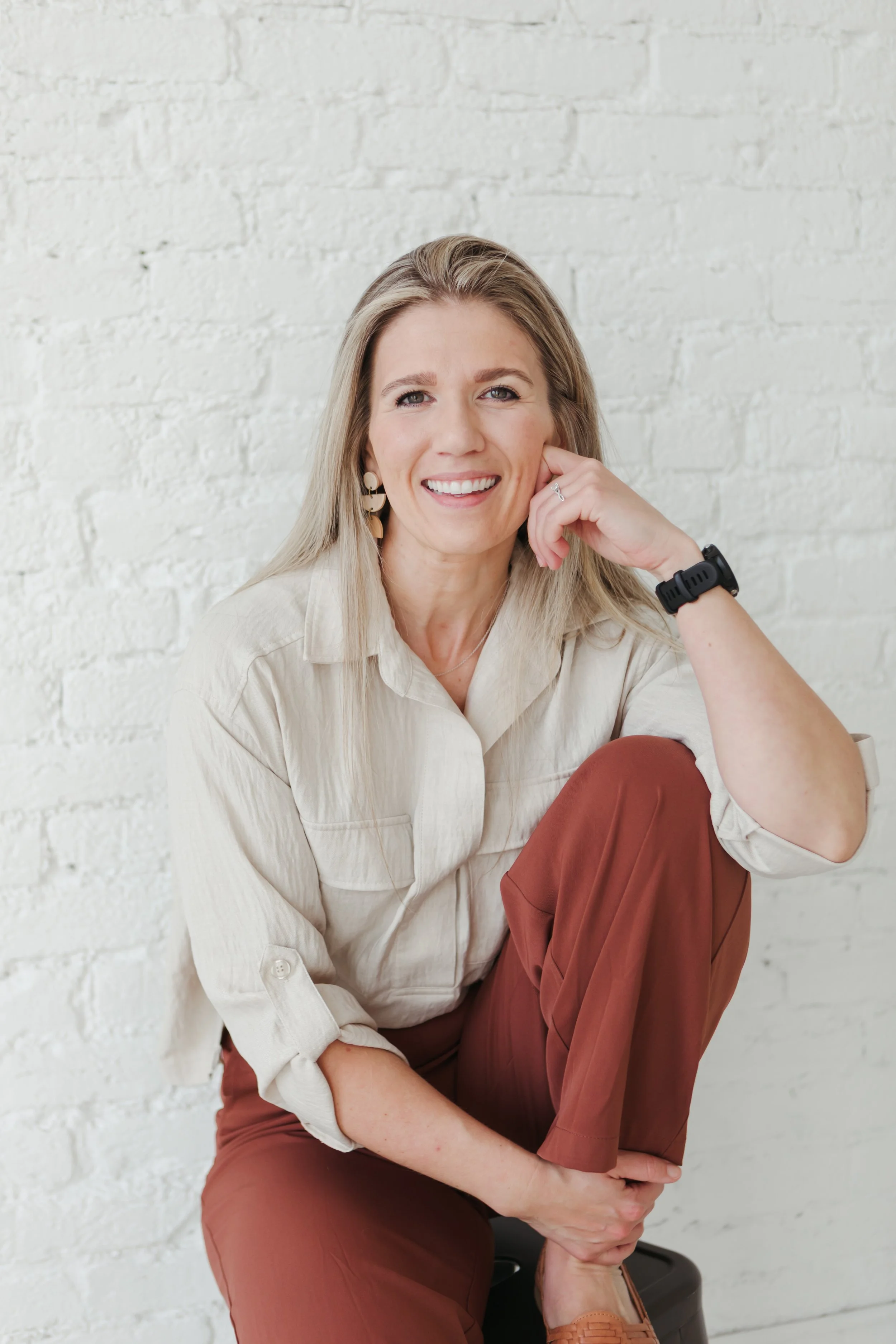 A woman with blonde hair, wearing earrings, a beige shirt, brown pants, and brown shoes, sitting against a white brick wall, smiling, with her left hand resting on her face and her right arm around her knee.