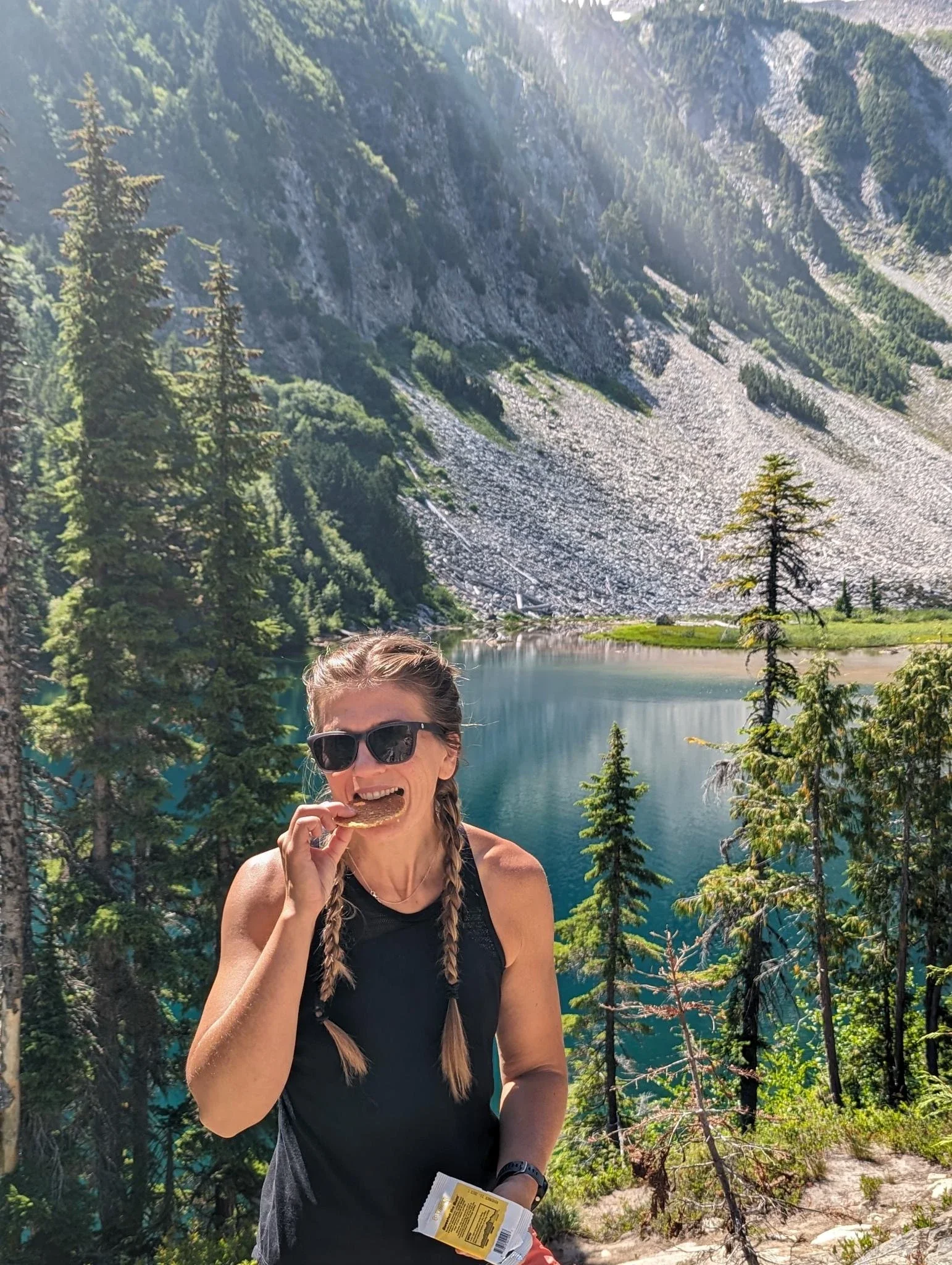 A woman with braided hair and sunglasses standing by a lake in a mountainous forest, eating a snack, with trees and a rocky mountain slope in the background.