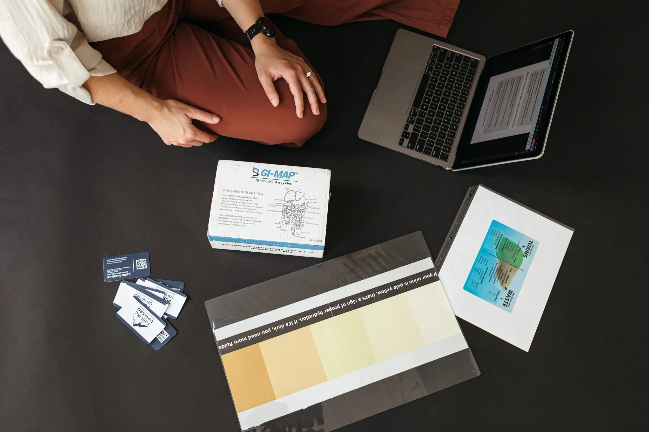 A person wearing a white shirt and brown pants sitting at a table with medical informational materials, including a box labeled GI-MAP DNA qPCR Stool Analysis, a color palette sample, a VASc score card, and a partially visible laptop and flight ticket.