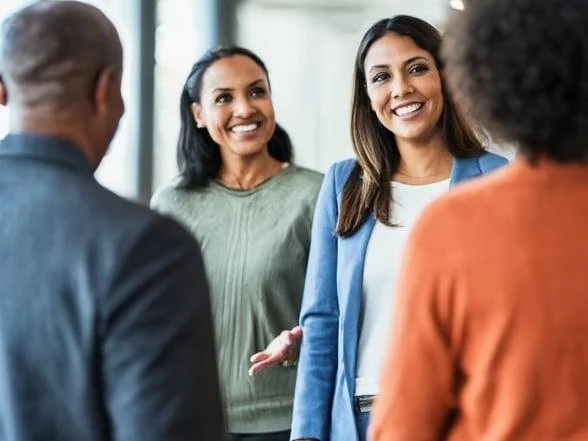 Four diverse people engaged in a friendly conversation indoors, smiling and gesturing.