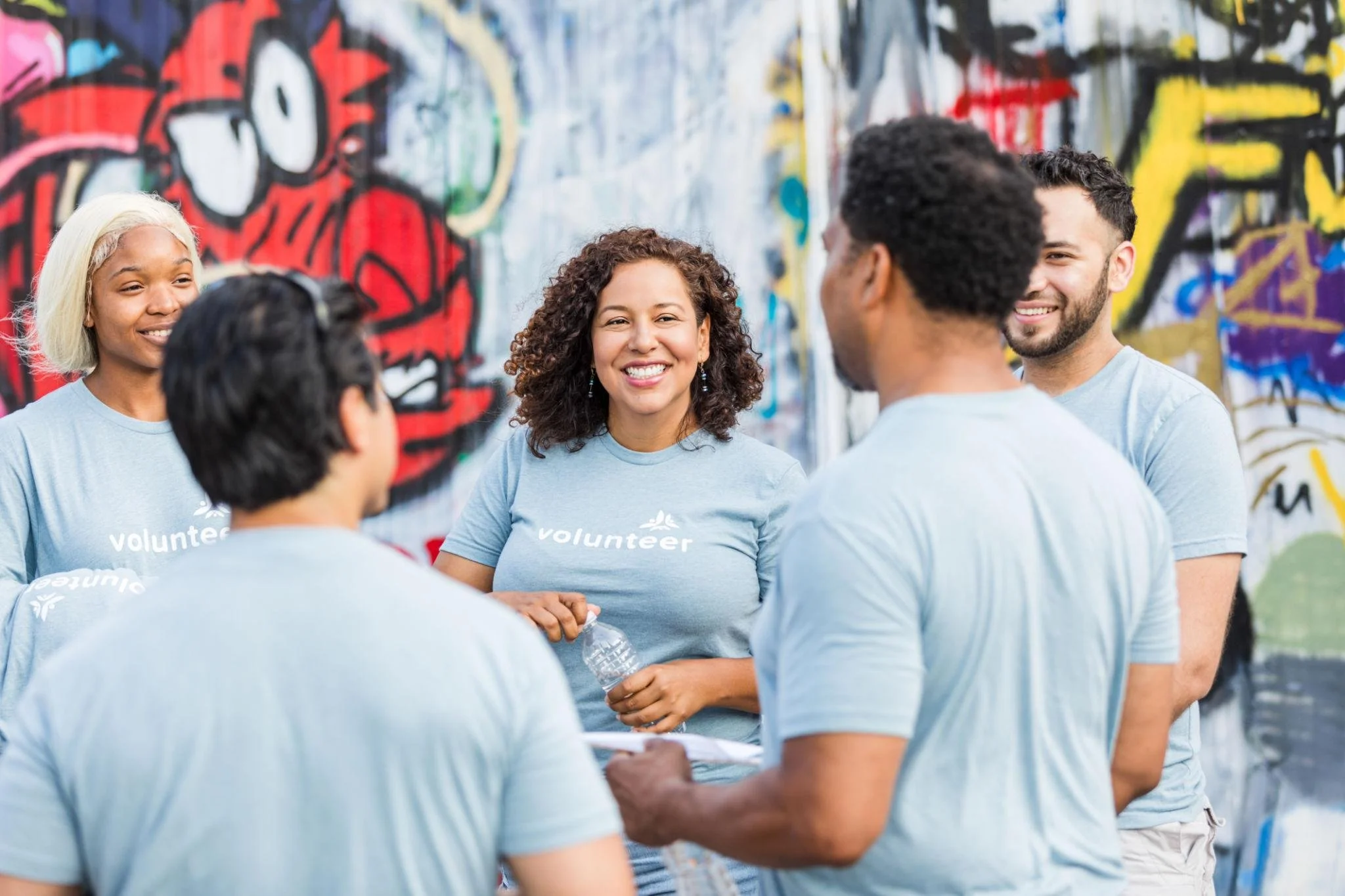 Group of diverse volunteers wearing light blue shirts with 'volunteer' printed on them, standing in front of a colorful graffiti wall, shaking hands, and smiling.