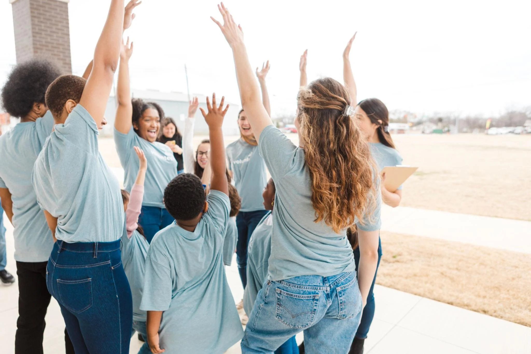 A group of diverse children and teenagers wearing light blue T-shirts, engaging in a group high five outdoors, with smiles and cheerful expressions.