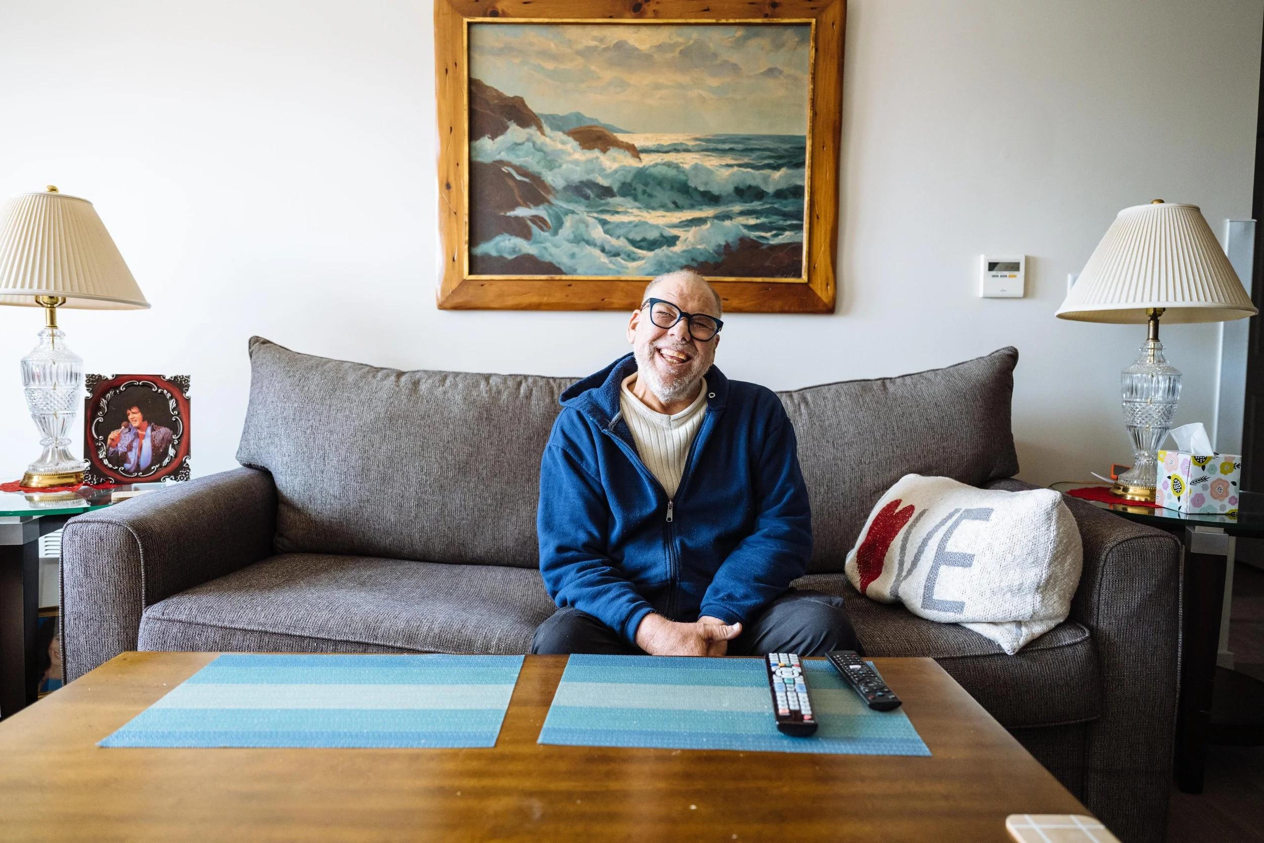 A man with glasses and a beard, smiling, sitting on a gray sofa in a living room. Behind him, a large painting of ocean waves and rocky cliffs hangs on the wall. There are two table lamps with pleated shades on side tables, and a framed picture of a singer on the left. In front of him, a wooden coffee table has place mats and remote controls.