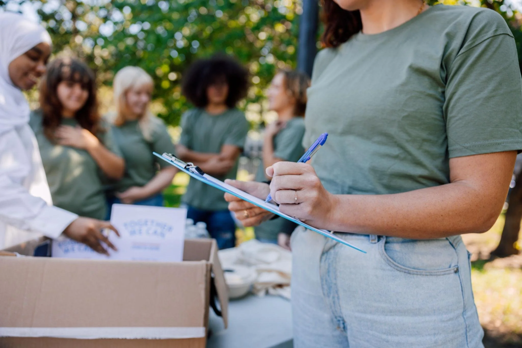 A woman in a green t-shirt and blue jeans is writing on a clipboard with a blue pen outdoors, with a group of women in casual clothing standing behind a table in a park with trees and sunlight.