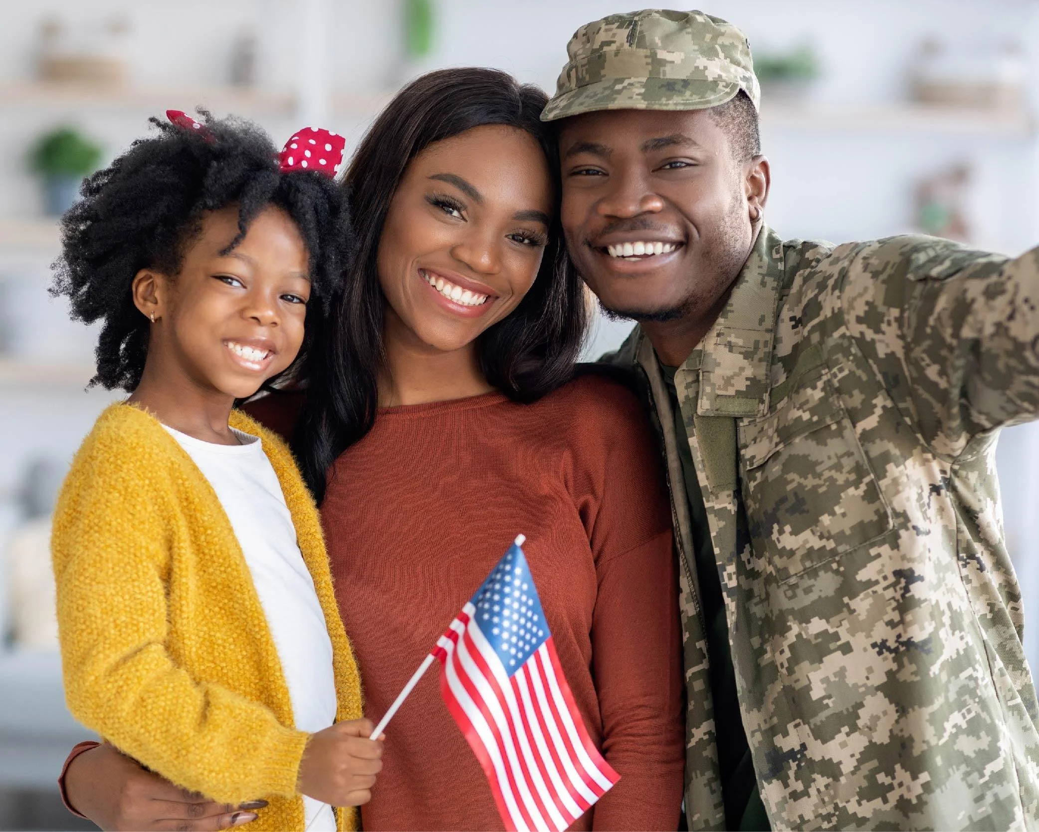 A smiling African-American woman, a young girl, and a man in military uniform taking a selfie together. The girl is holding American flags and wearing a yellow cardigan and a red polka-dot bow in her hair. The woman is wearing a maroon top, and the man is in camouflage uniform with a cap. They appear to be celebrating an American holiday or event.