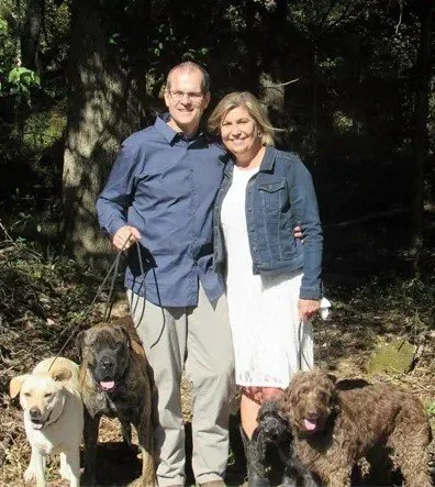 Doug and Karen Oler standing outdoors with four dogs, with trees in the background.