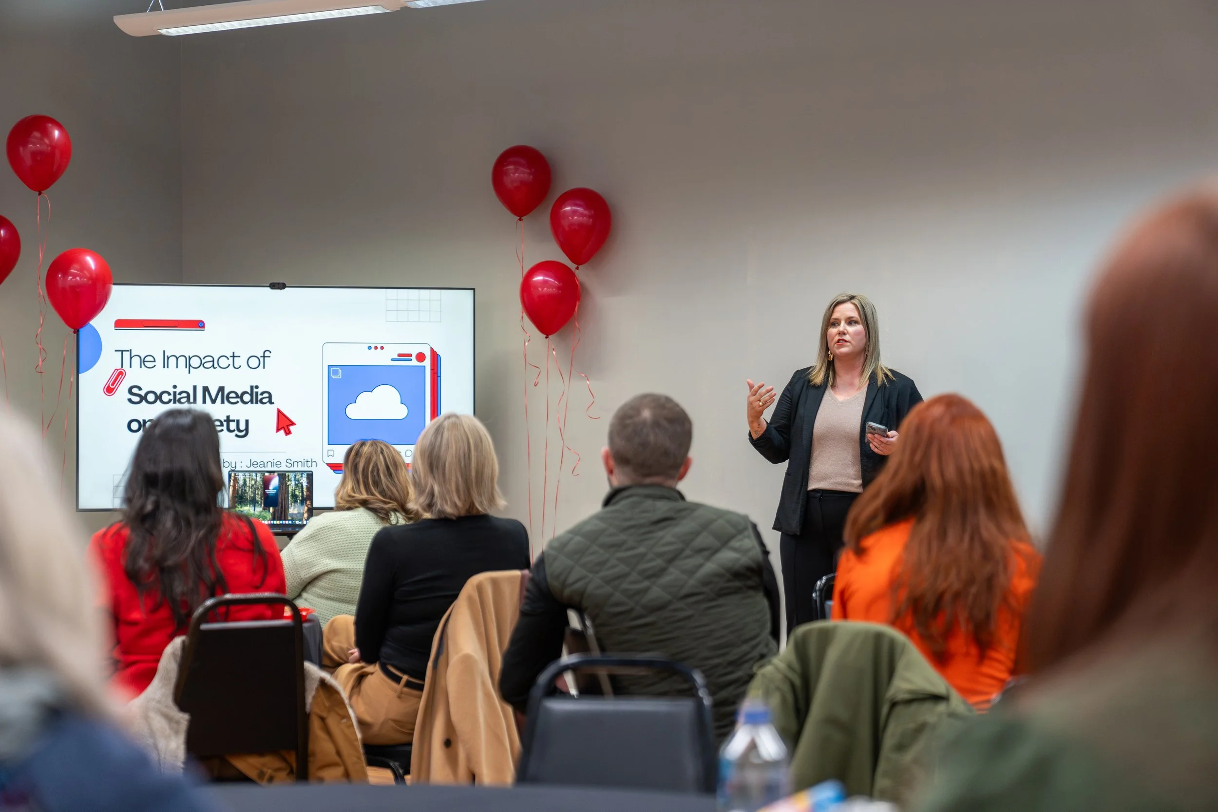 A woman in a black jacket and beige top giving a presentation to an audience about social media and security, with red balloons and a large screen display in a conference room.