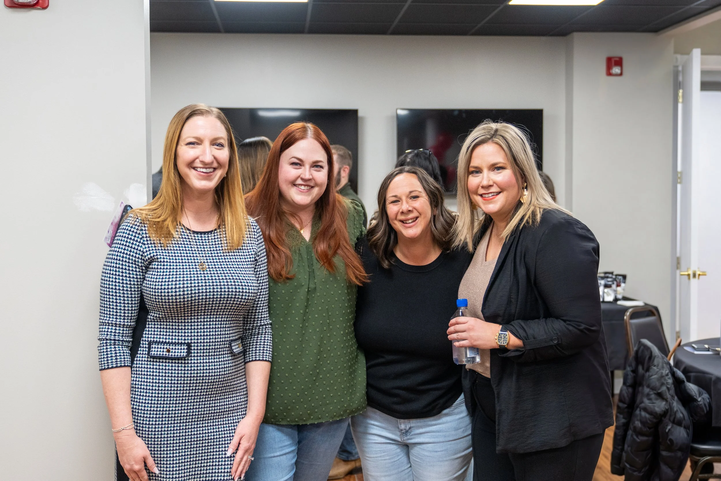 Four women smiling and standing together indoors at a social gathering, with a few other people and television screens in the background.