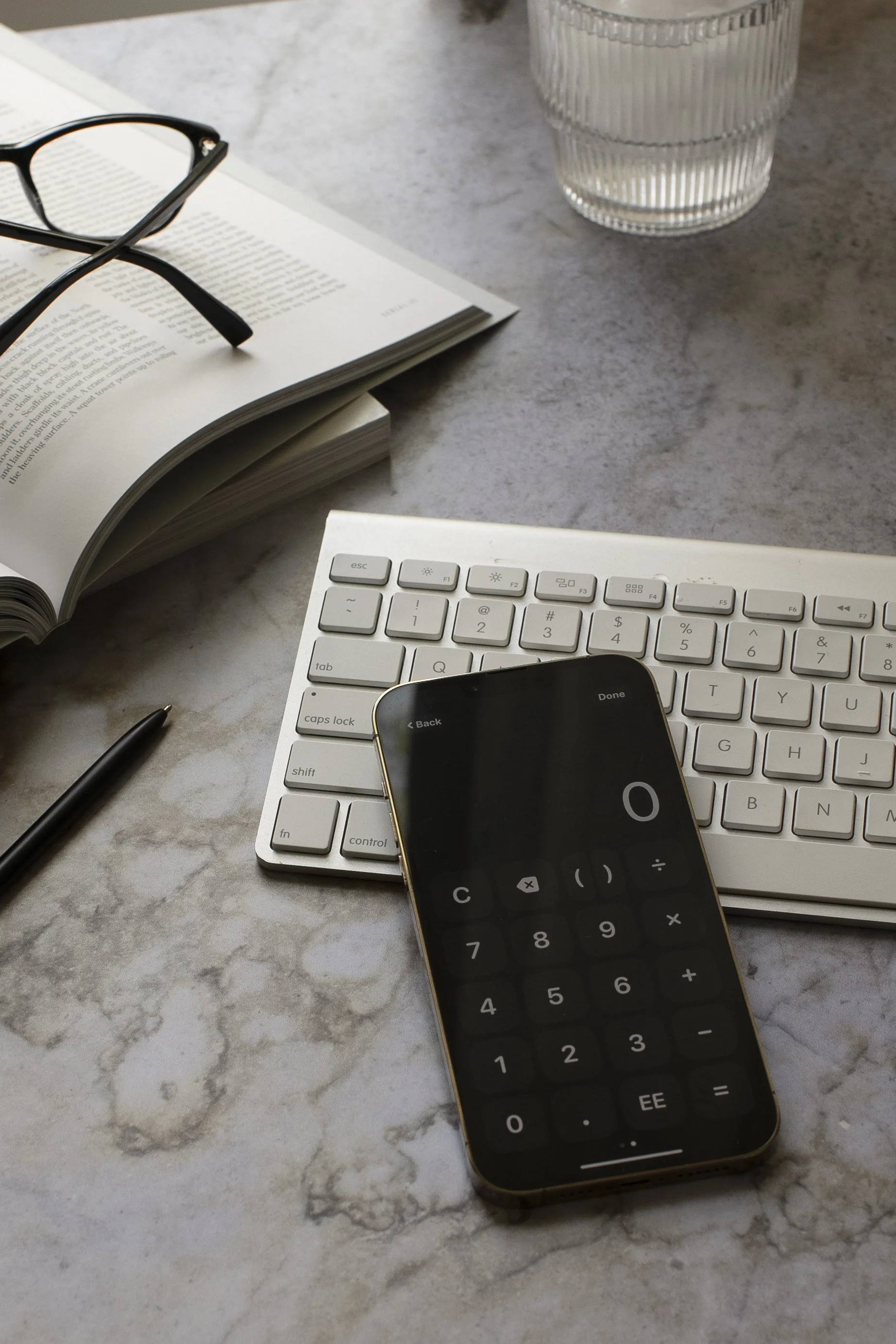 A workspace on a marble table featuring a computer keyboard, a smartphone with a calculator app open, an open book with glasses resting on it, a black pen, and a glass of water.