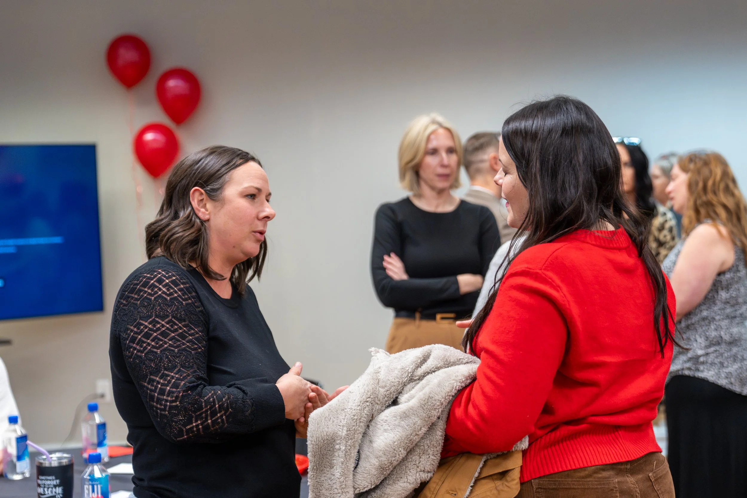 Two women are engaged in a handshake and conversation at a social event, with other people in the background. Red balloons are visible on the wall behind them.